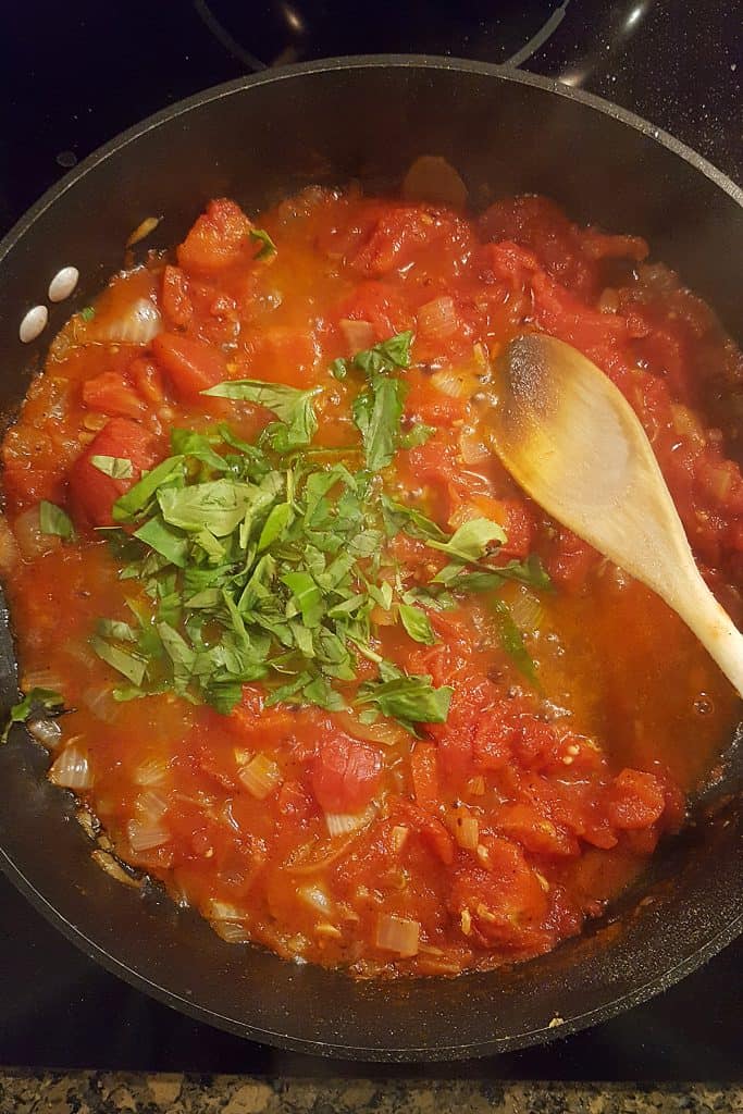 A skillet contains a tomato sauce with chopped onions and a pile of fresh chopped basil on top. A wooden spoon rests in the sauce, and the mixture is being cooked on a stovetop.