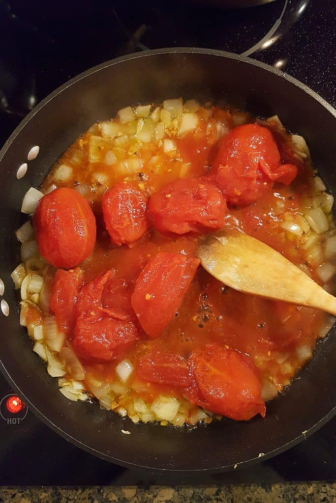 A skillet on a stove contains chopped onions and whole peeled tomatoes being cooked together. A wooden spoon rests in the pan, and the stove’s burner is set to hot.