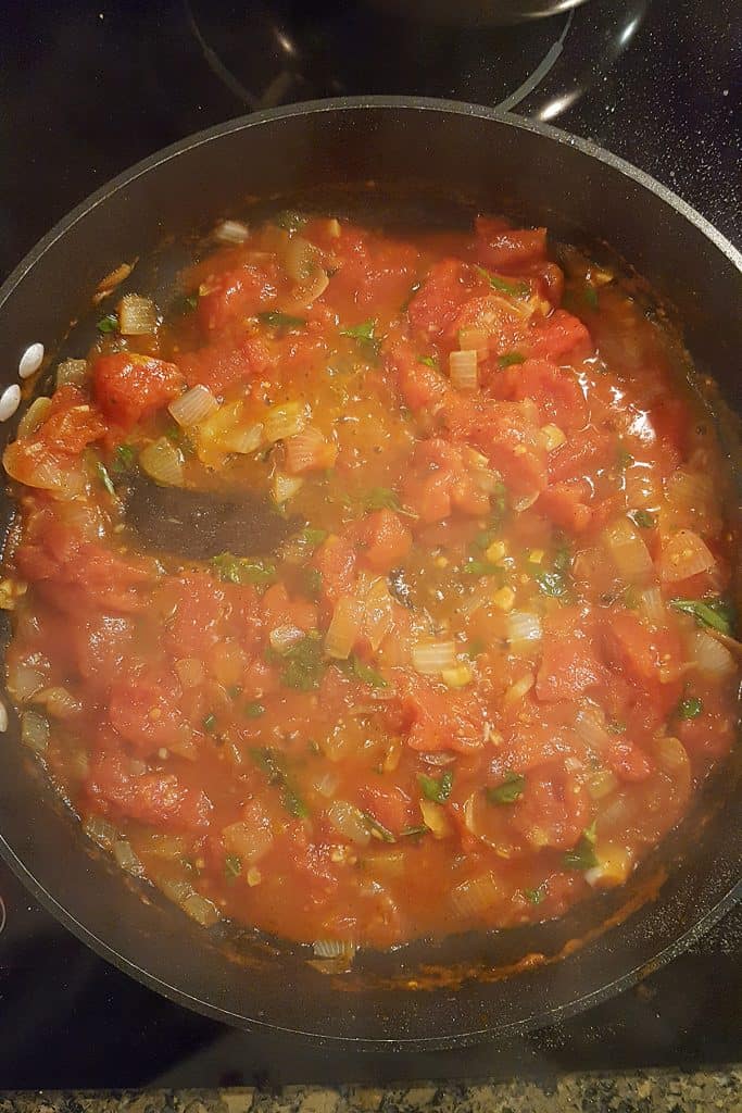 A pan filled with a tomato-based sauce containing chopped tomatoes, onions, herbs, and visible cooking liquid, being stirred with a black spoon on a stovetop.