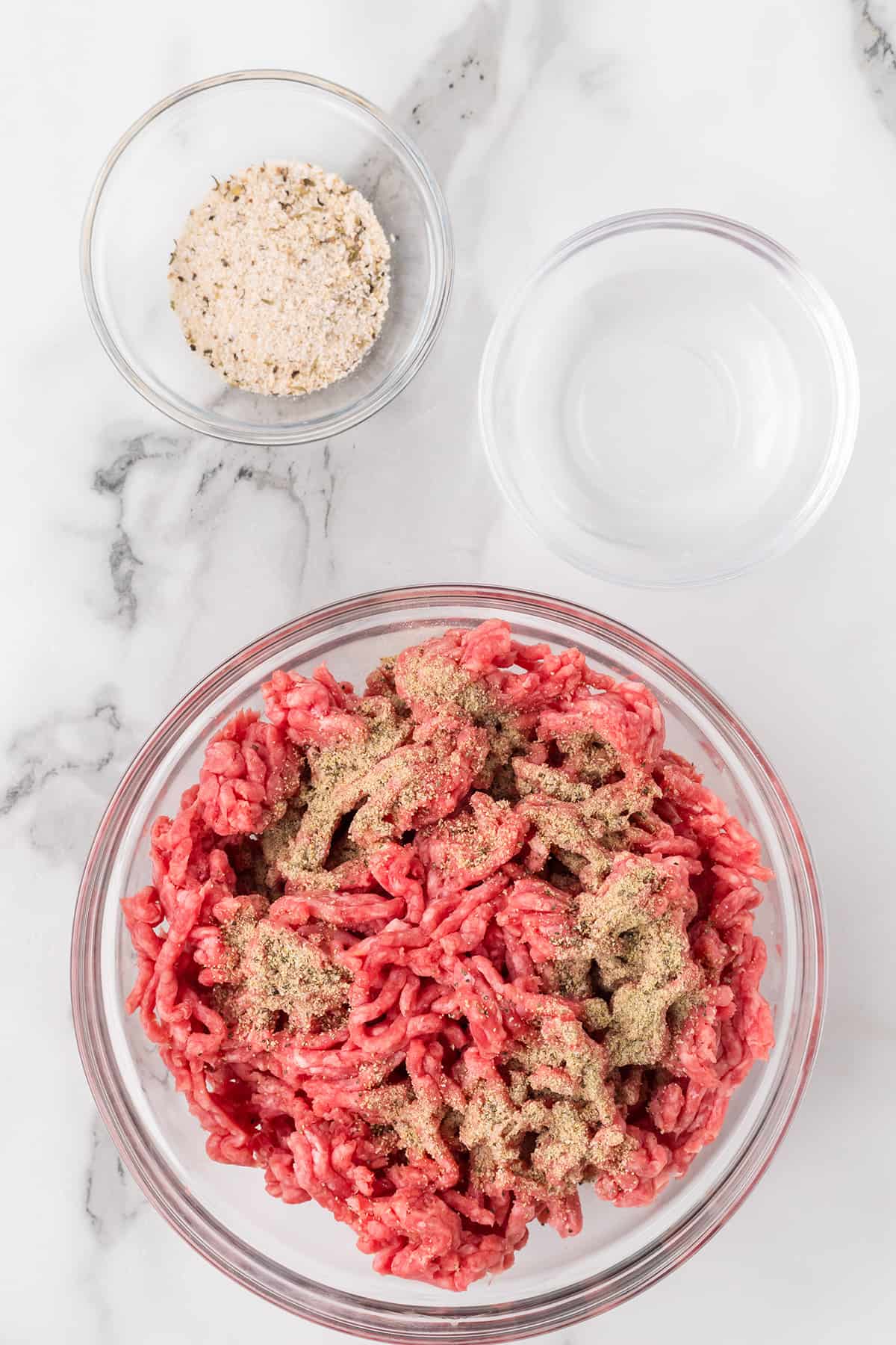 Ground beef with seasonings in a bowl, with additional seasonings and water in separate small bowls on a marble surface.