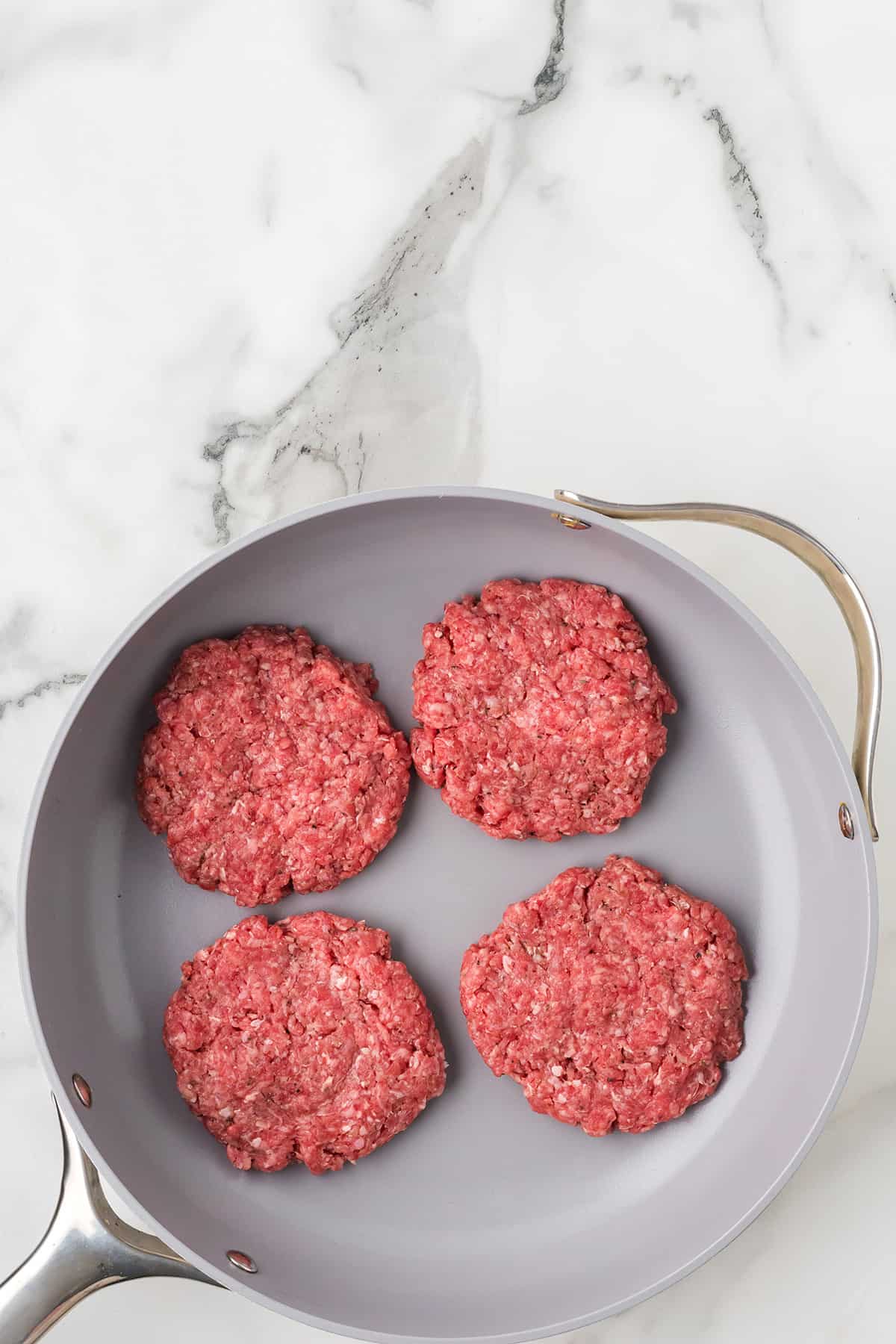 Four raw ground beef patties arranged in a nonstick frying pan on a white marble countertop.