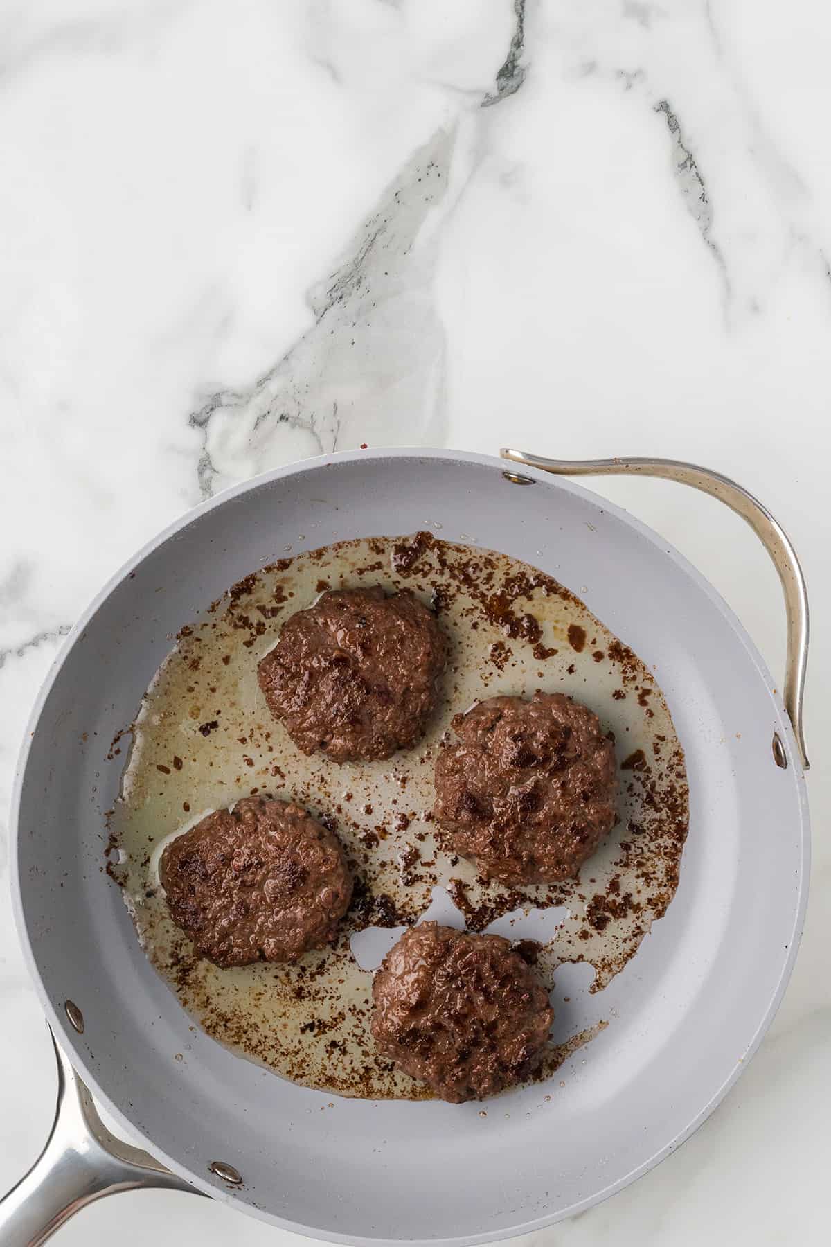 Four cooked beef patties in a frying pan with browned residue on a white marble surface.