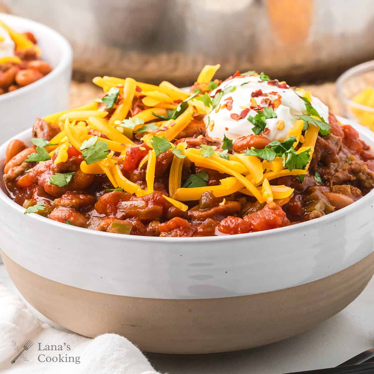 A bowl of chili topped with shredded cheese, sour cream, chopped cilantro, and red pepper flakes.