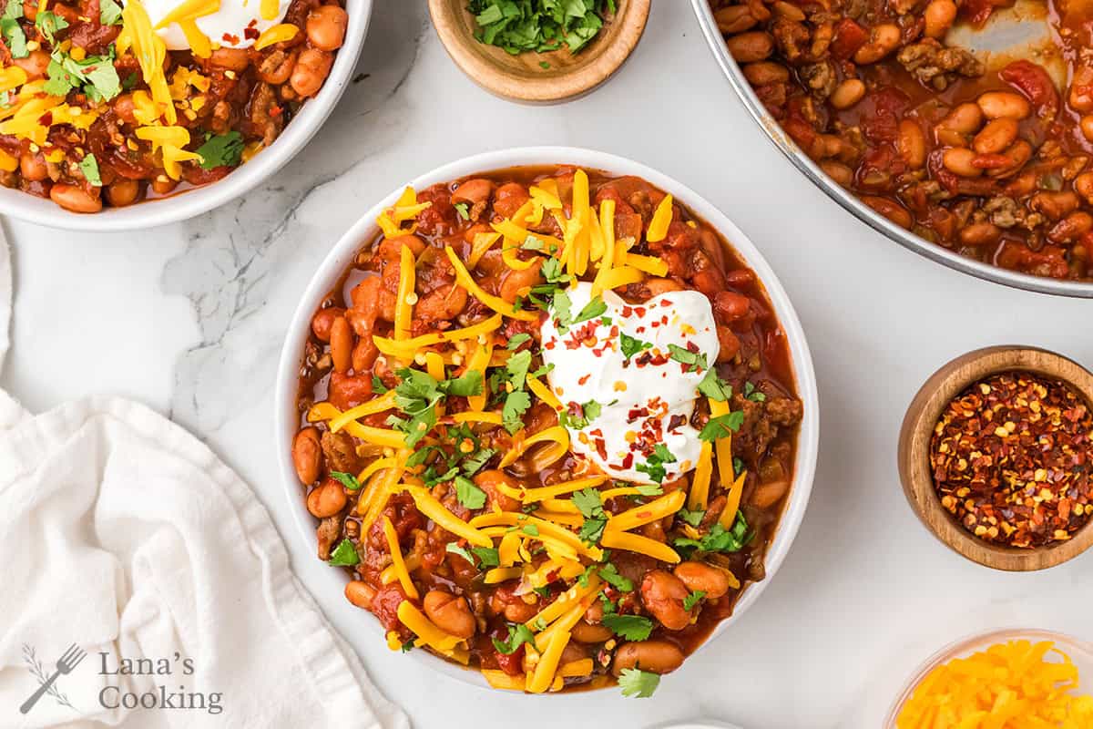 A bowl of chili topped with shredded cheese, sour cream, cilantro, and red pepper flakes on a white surface.