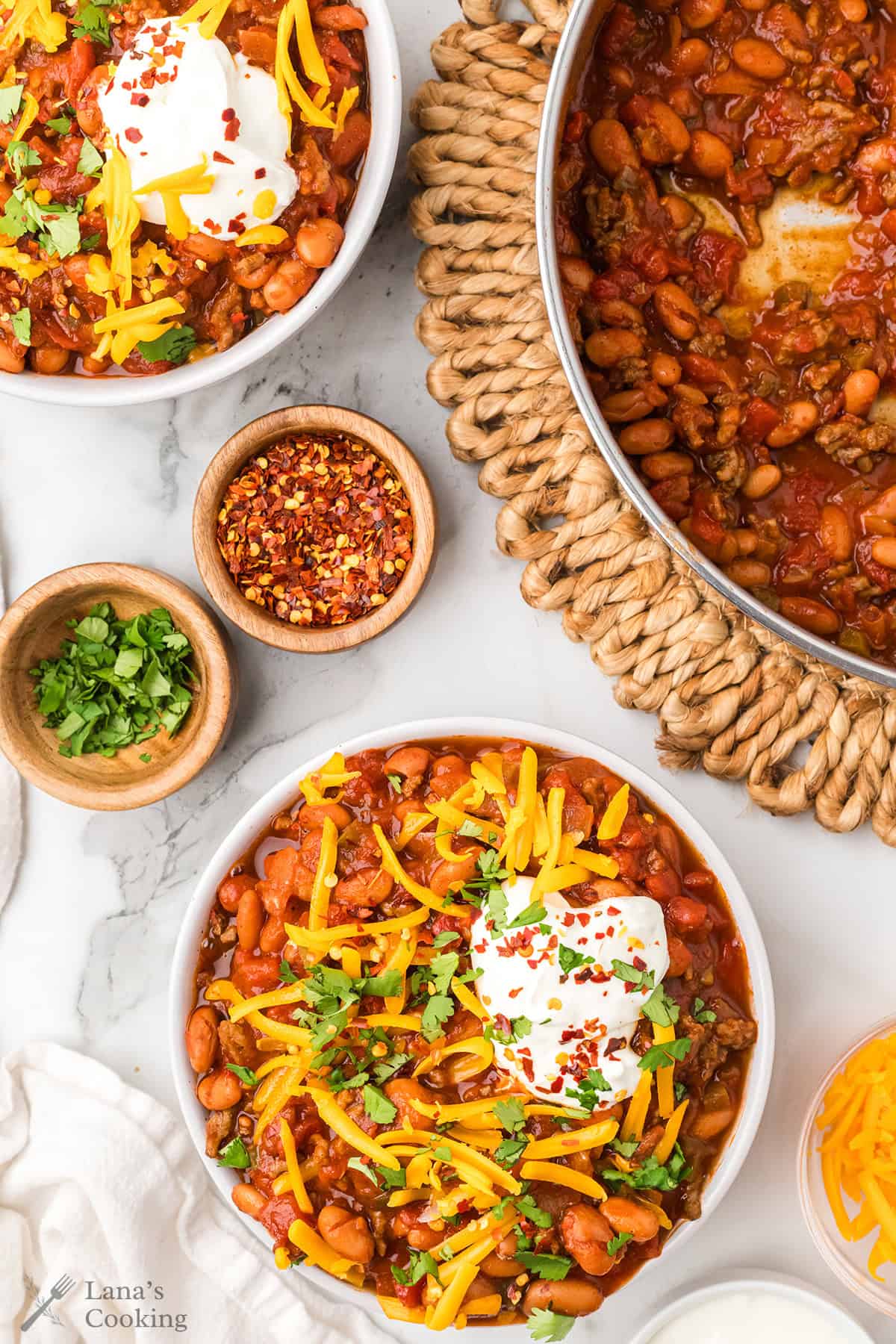 Bowls of chili topped with shredded cheese, sour cream, cilantro, and red pepper flakes, with garnishes on the side.