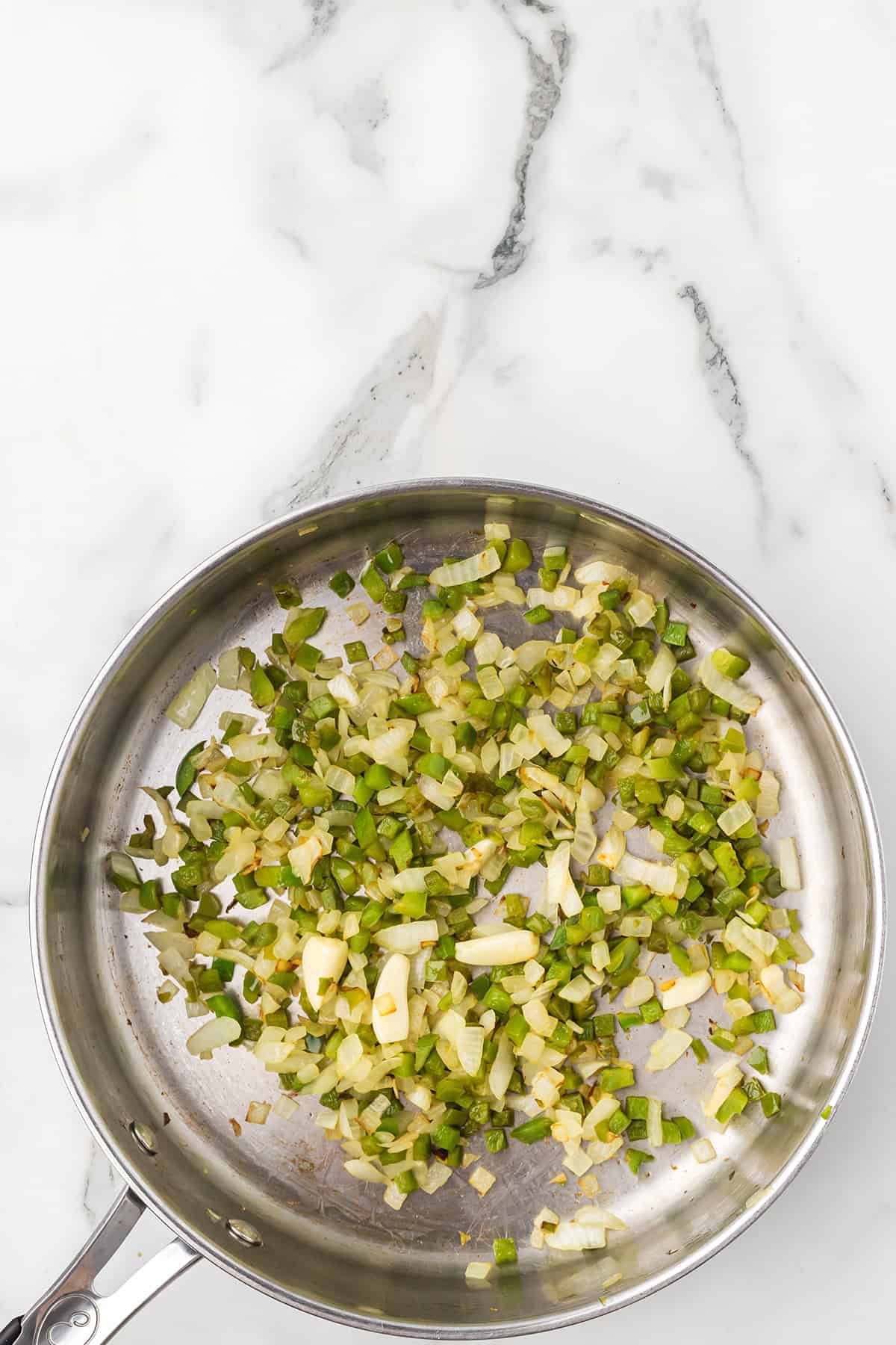 Chopped onions and green peppers sautéing in a stainless steel pan on a marble countertop.
