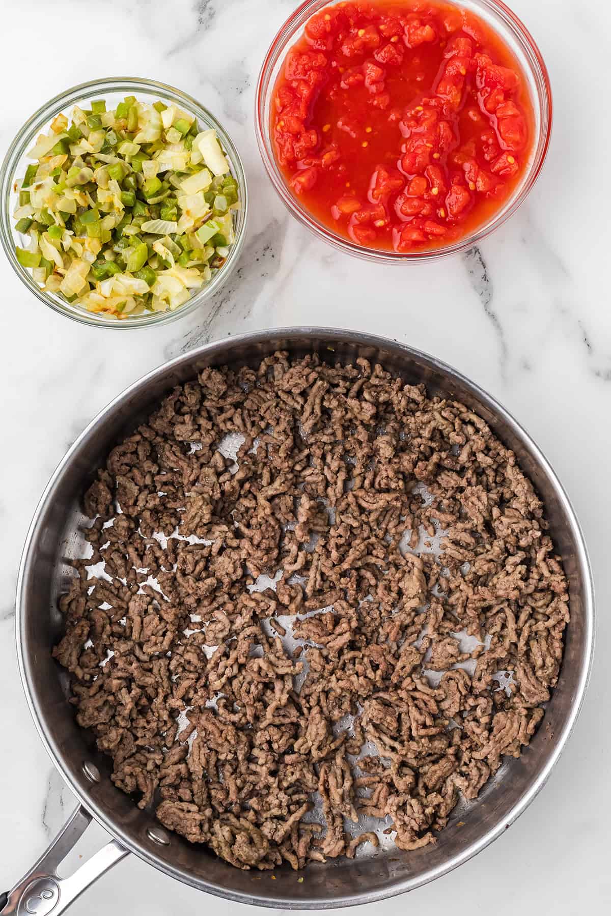 Cooked ground beef in a skillet next to bowls of chopped vegetables and diced tomatoes on a marble counter.