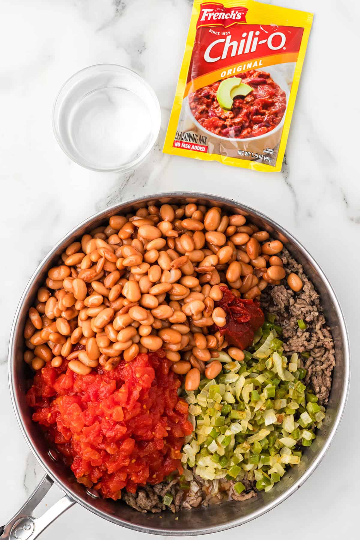 A skillet with beans, ground beef, tomatoes, green chilies, and chili seasoning packet on a marble counter.