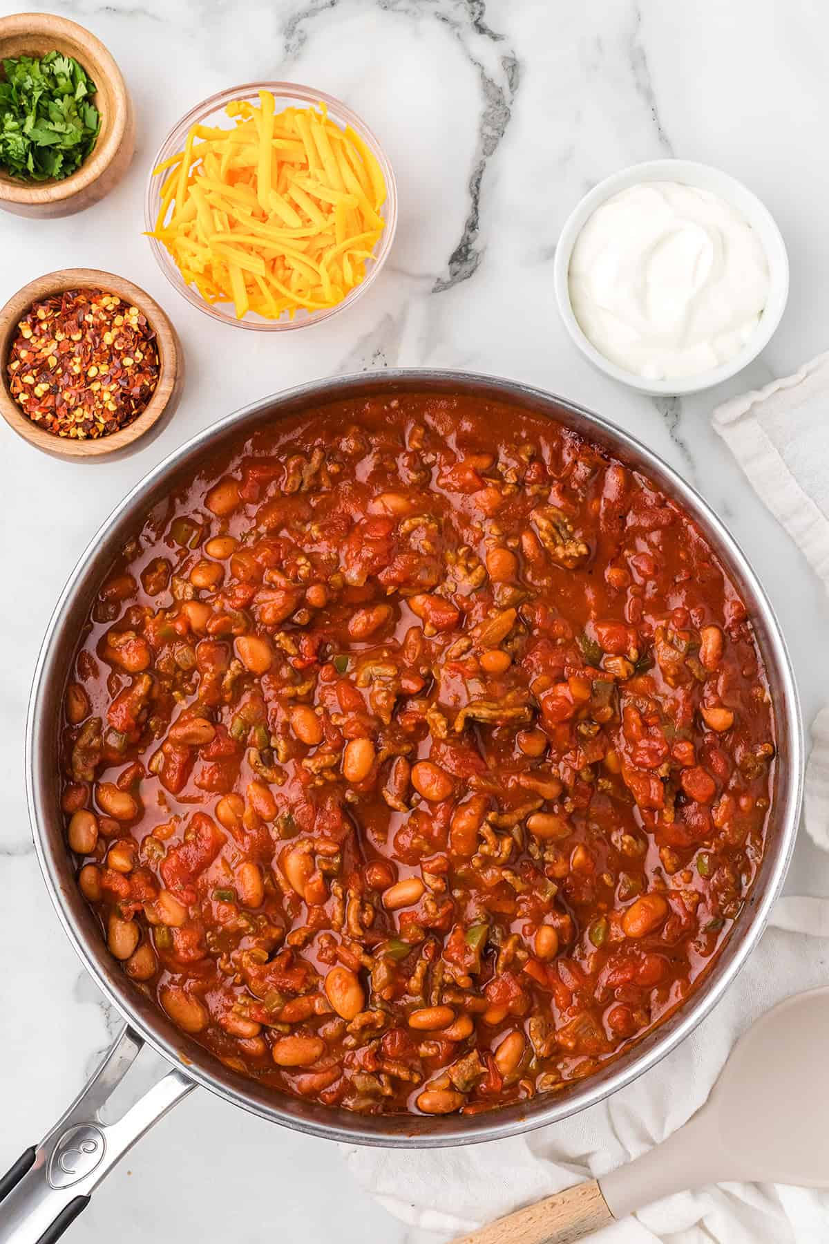 A pan of chili with beans and tomatoes, surrounded by bowls of shredded cheese, sour cream, herbs, and chili flakes.