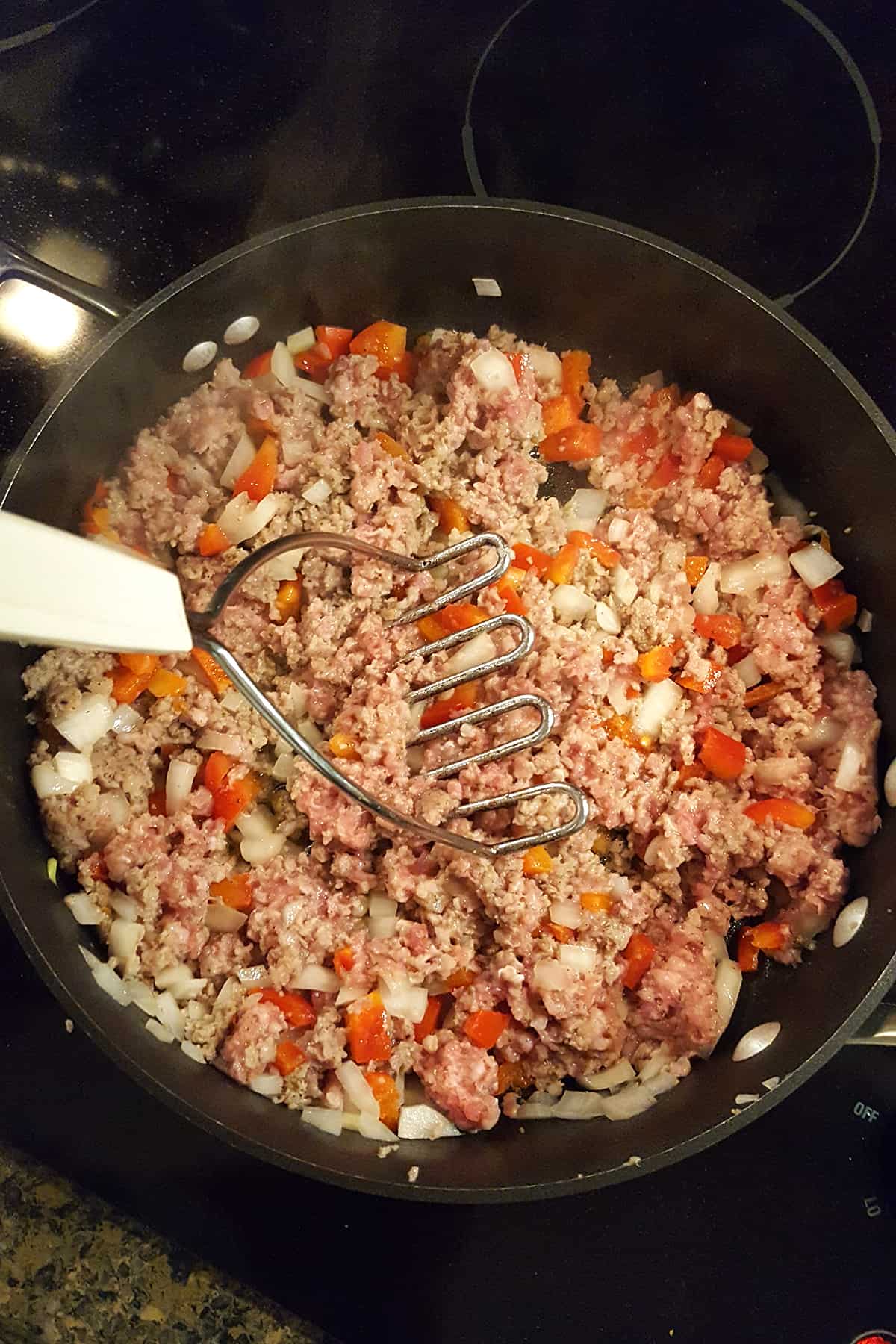 Onions, peppers, and sausage in a skillet with a potato masher breaking up the sausage meat.