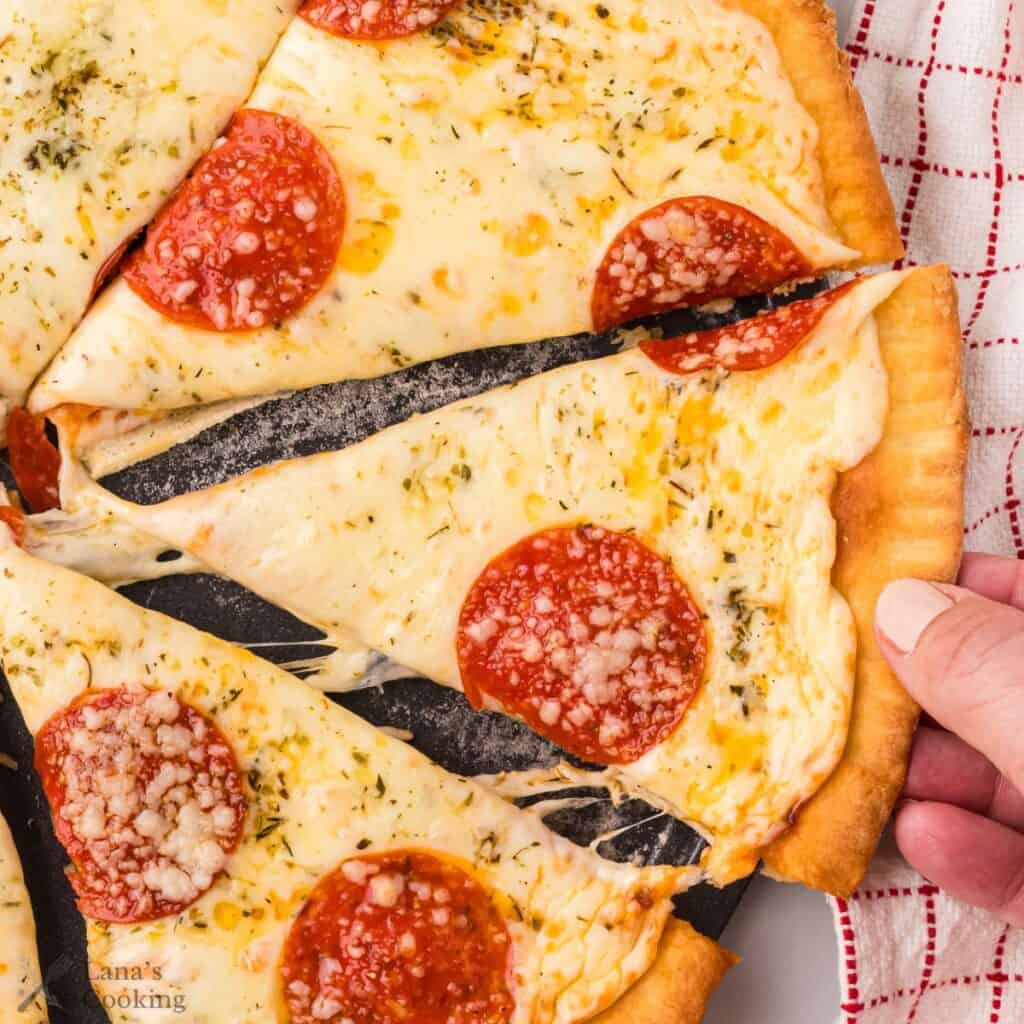 A hand lifts a slice of pepperoni pizza with melted cheese from a pizza on a dark tray, next to a red and white towel.