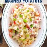 Oval dish of mashed potatoes with bacon, tomato, and green onions, on a wooden table with a spoon beside it.