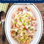 Oval dish of mashed potatoes mixed with bacon, tomatoes, and green onions, next to a spoon and green napkin.