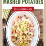 Oval dish of mashed potatoes with bacon and tomatoes, garnished with green onions, on a wooden table.