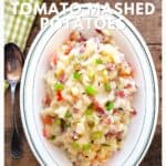 Oval dish of mashed potatoes with bacon, tomatoes, and green onions, beside a spoon and checkered napkin.