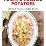 Bowl of mashed potatoes with bacon, tomato, and green onions on a wooden table, spoon and napkin beside.