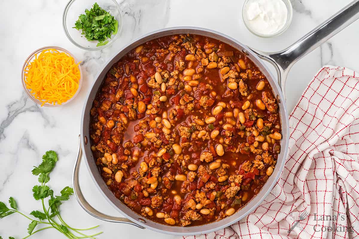A pot of chili with ground meat and beans, surrounded by shredded cheese, cilantro, and sour cream in bowls.