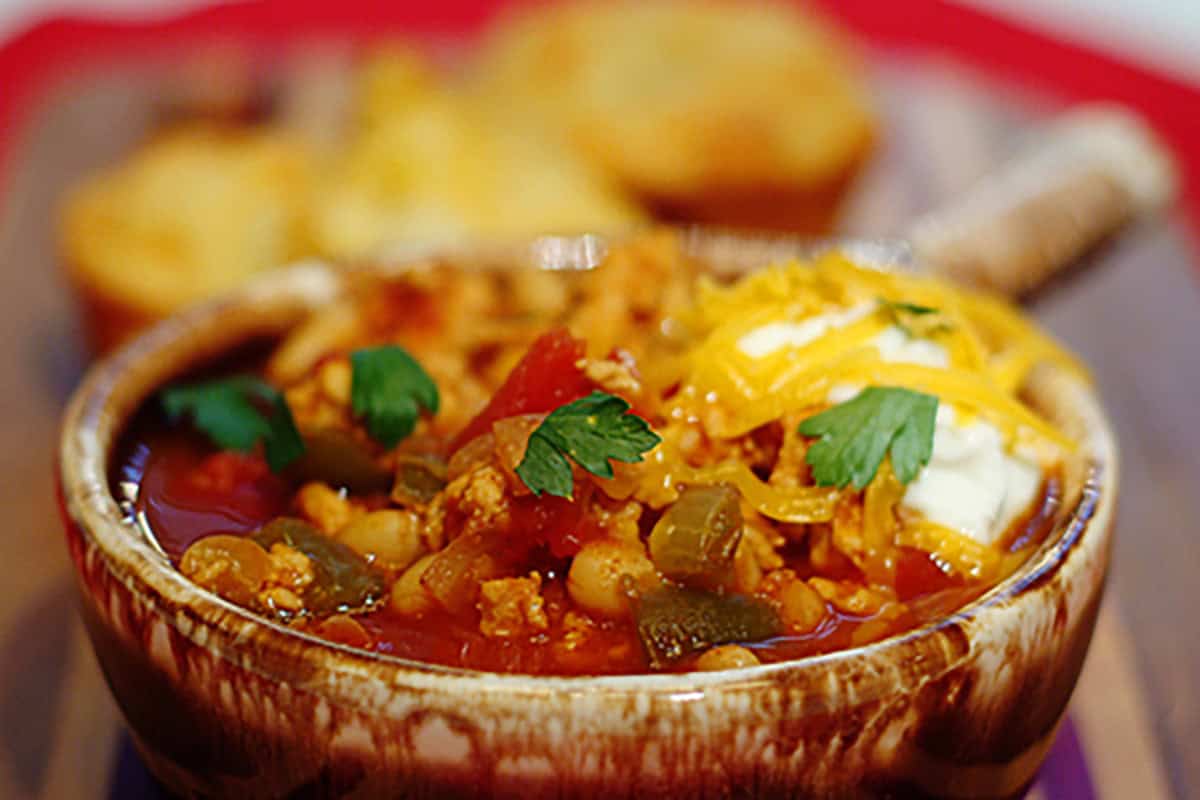 A bowl of chili topped with shredded cheese, sour cream, and parsley, with cornbread in the background.