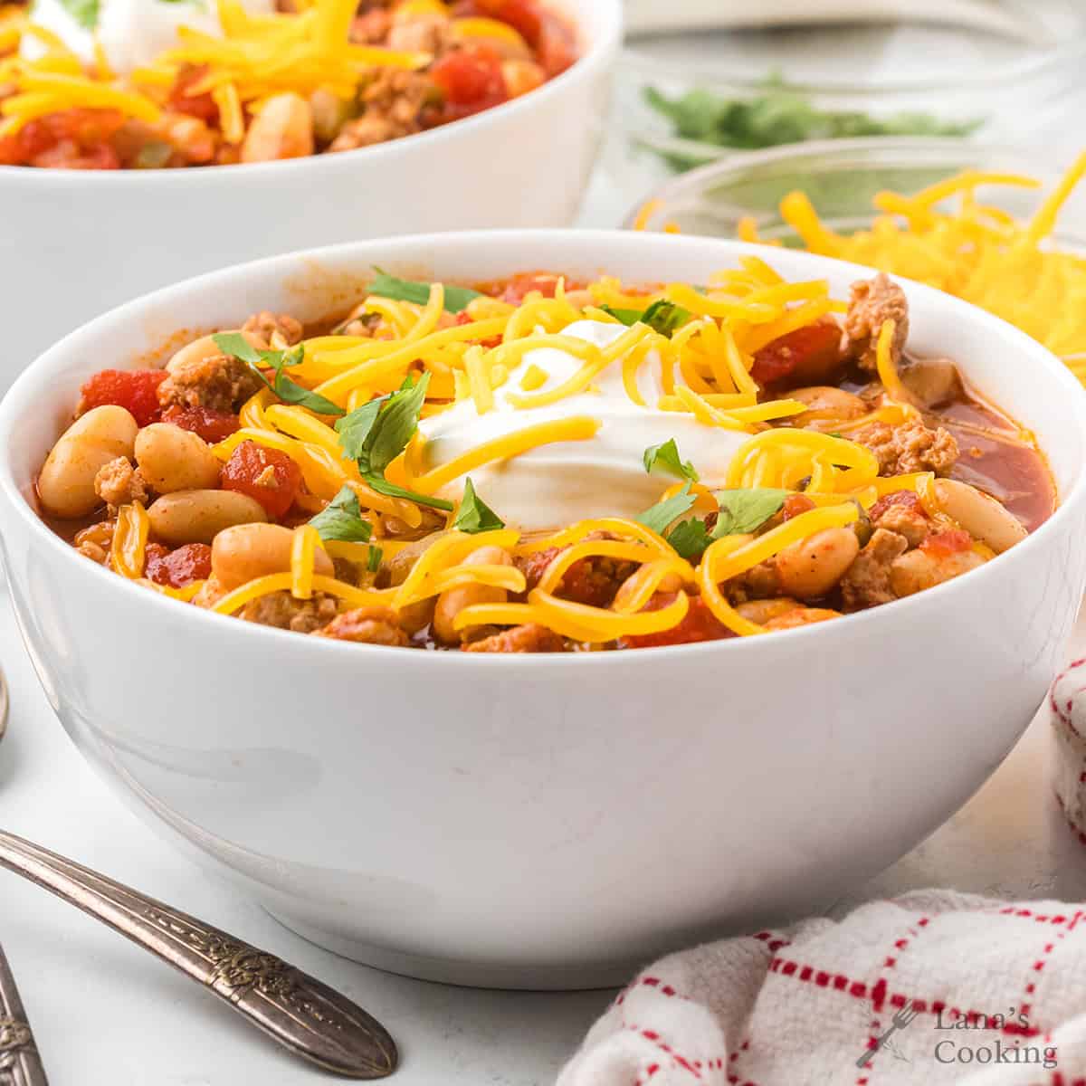 A bowl of chili topped with shredded cheese, sour cream, and fresh herbs, with a spoon and napkin beside it.