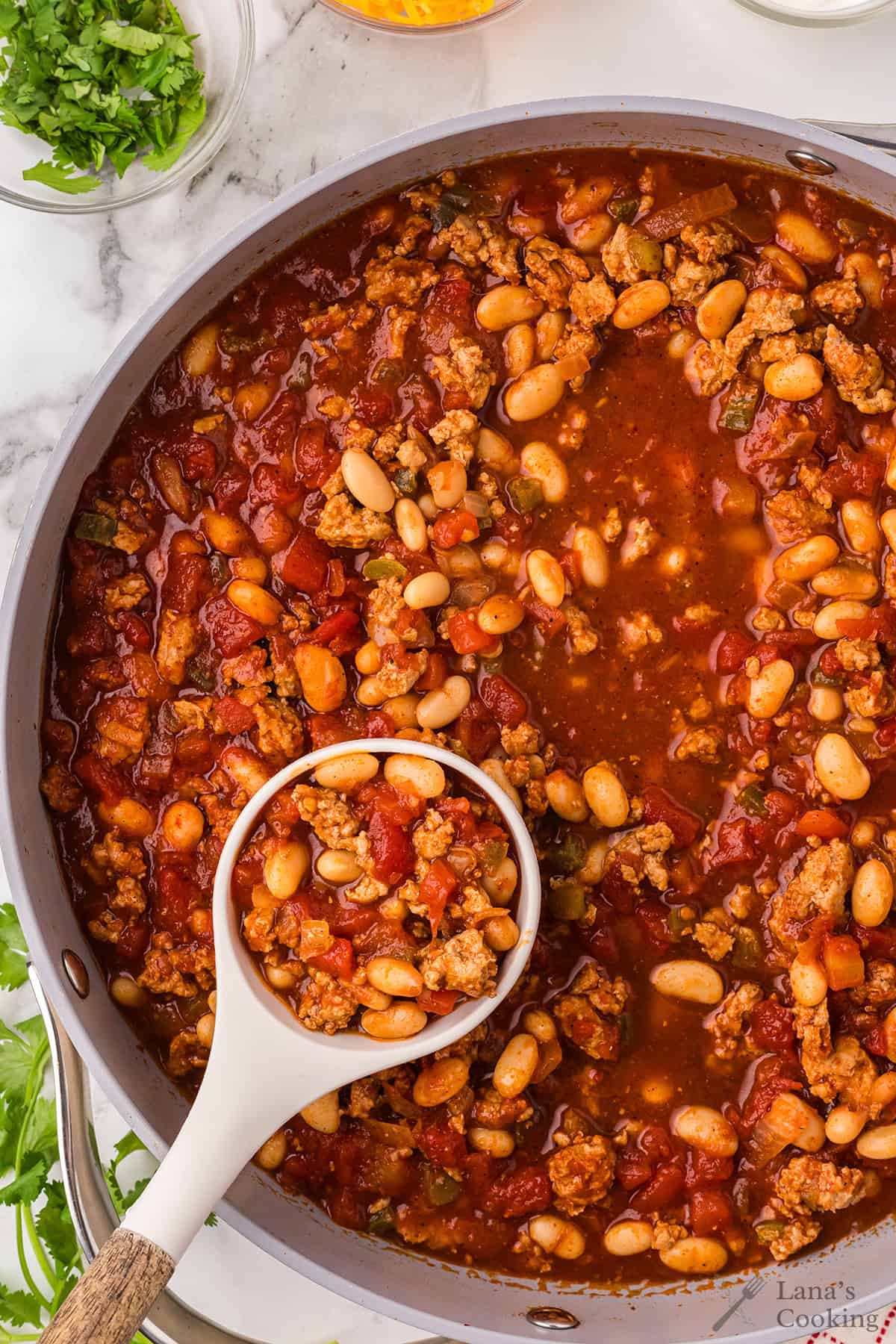 A pot of chili with ground meat, white beans, tomatoes, and a ladle scooping some out.