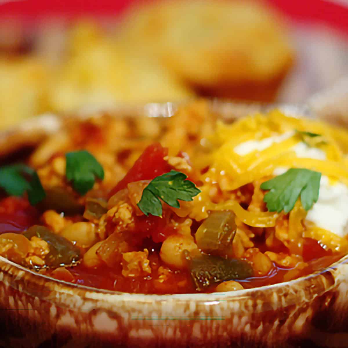 A bowl of chili topped with shredded cheese, parsley, and sour cream, with cornbread in the background.