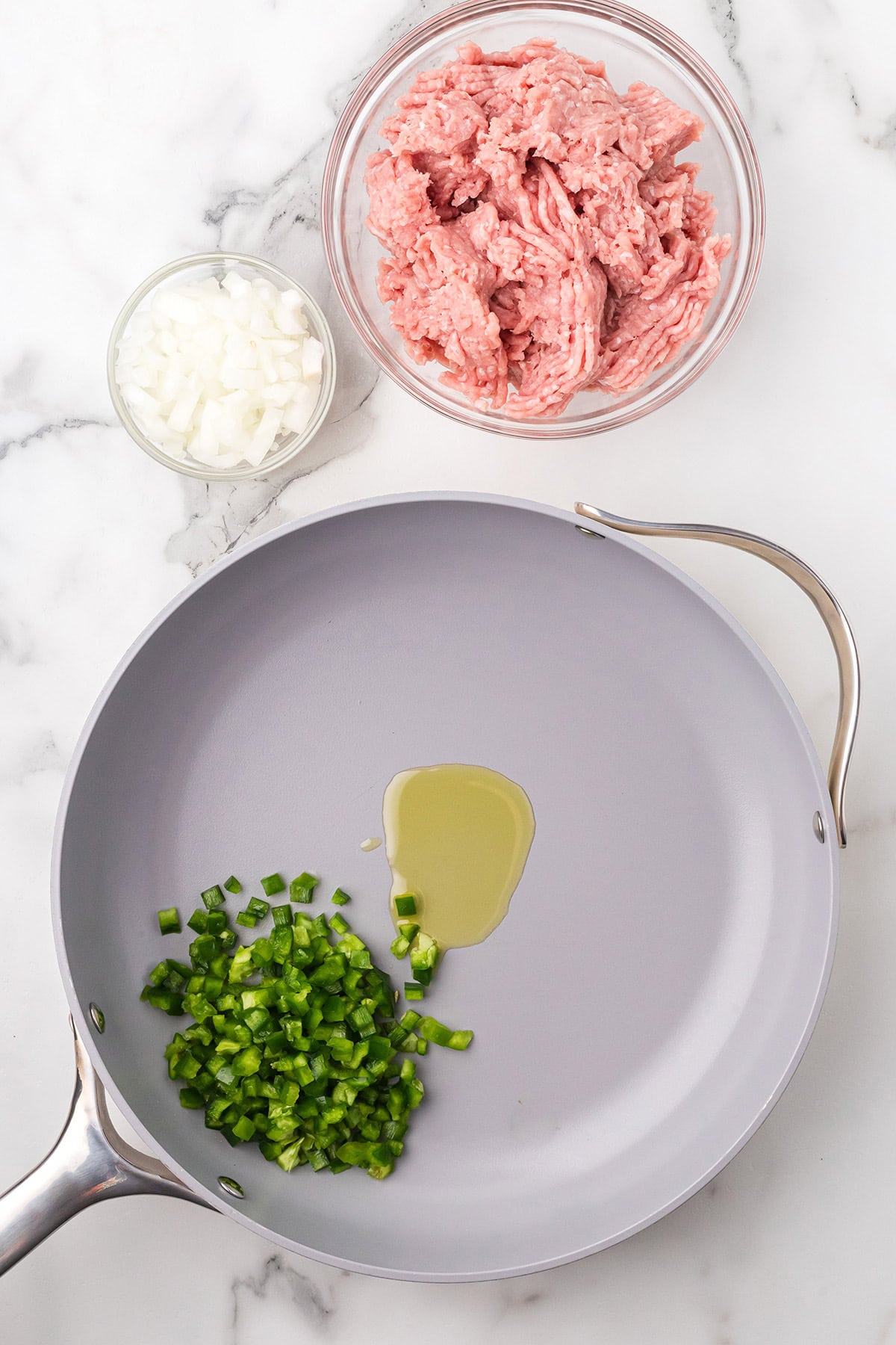 A pan with oil and chopped green pepper, next to bowls of chopped onion and raw ground meat on a marble counter.