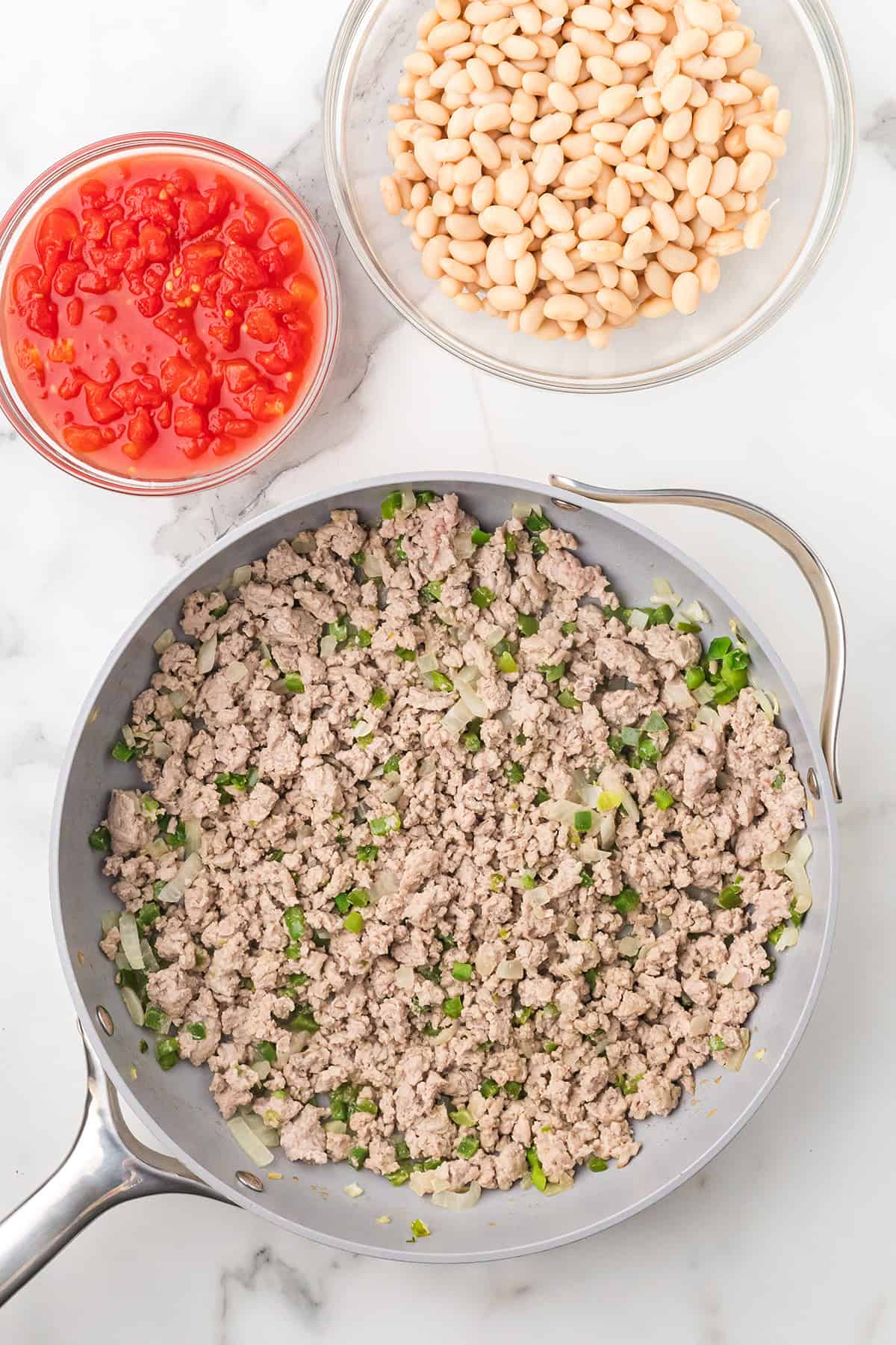 Cooked ground meat with onions and peppers in a skillet, next to bowls of diced tomatoes and white beans.