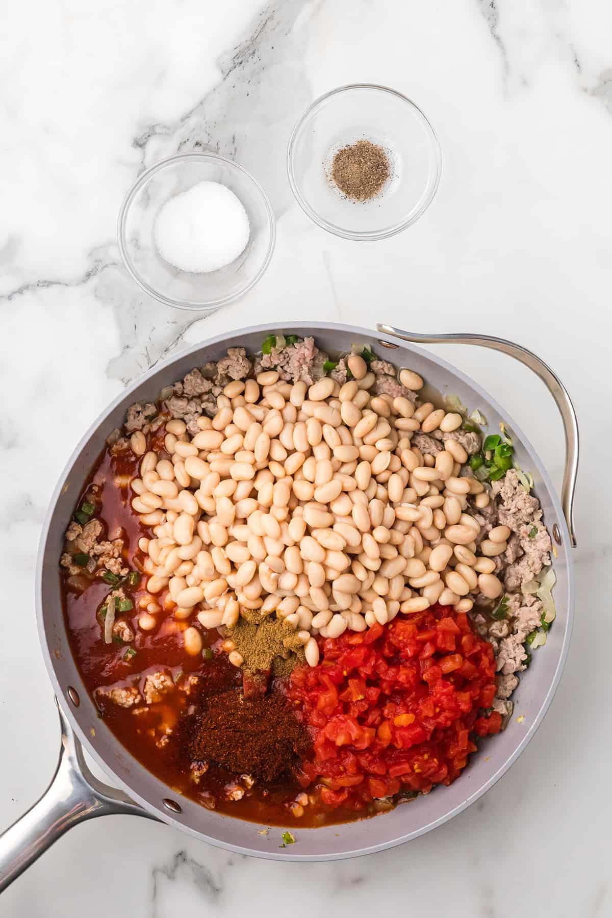 A skillet with ground meat, white beans, diced tomatoes, and spices, plus bowls of salt and pepper nearby.