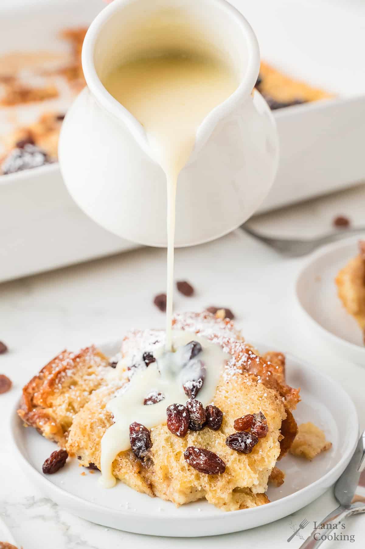 A close-up of a white pitcher pouring vanilla sauce over a serving of bread pudding with raisins on a white plate. The bread pudding is dusted with powdered sugar and more bread pudding is visible in the background.