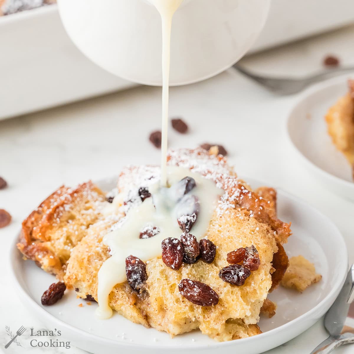 A serving of bread pudding topped with raisins is on a white plate, being drizzled with a creamy sauce. A fork is placed beside the plate, and another plate of bread pudding is visible in the background.
