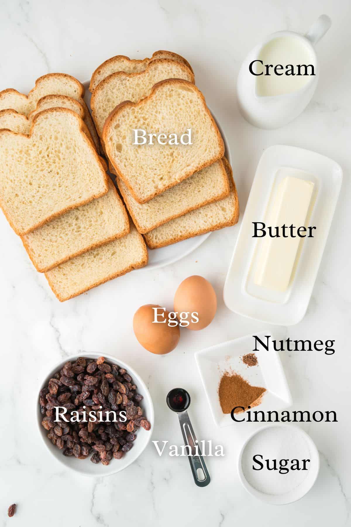 Slices of bread, a bowl of raisins, two eggs, a stick of butter on a dish, a small pitcher of cream, a dish of sugar, a measuring spoon of vanilla, and small piles of nutmeg and cinnamon on a white surface.