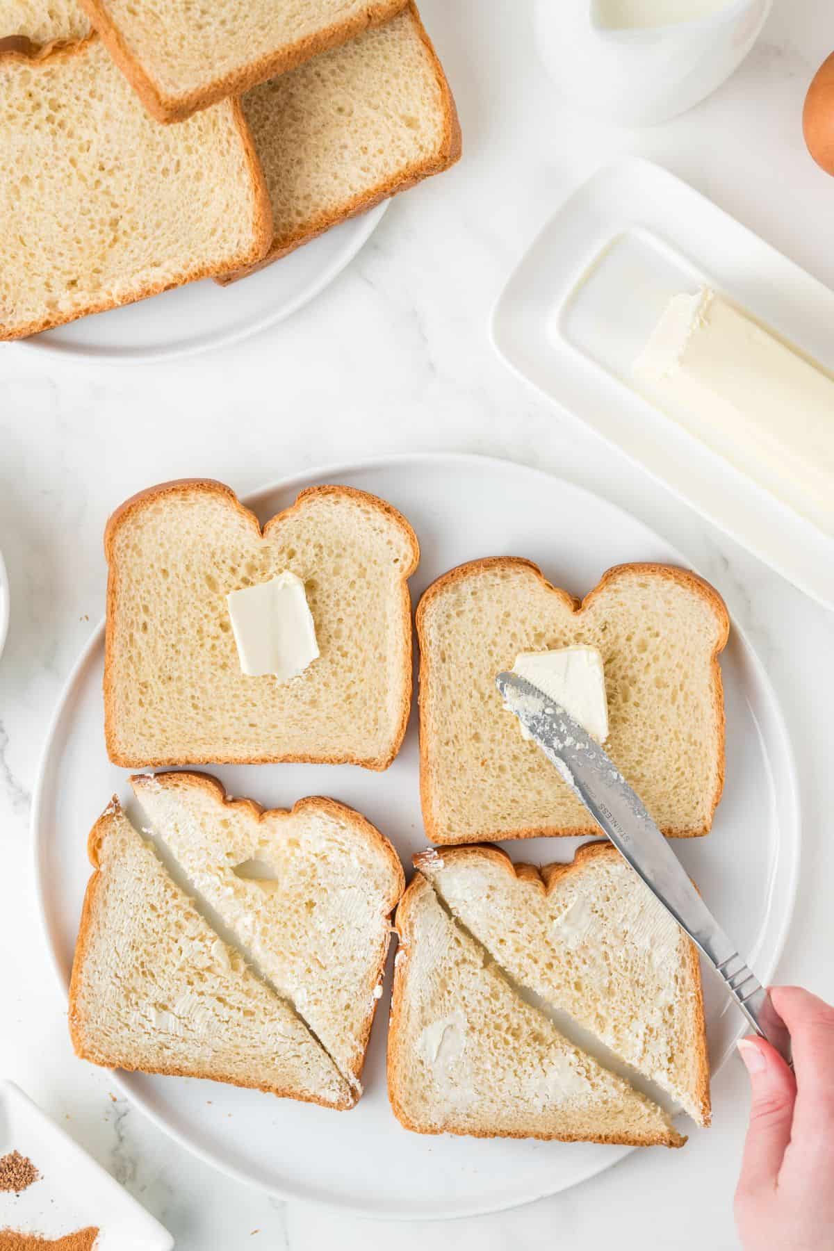 A plate with four slices of white bread, two with pats of butter, and two being spread with butter using a knife. More bread, a stick of butter, and a milk pitcher are nearby on a white surface.