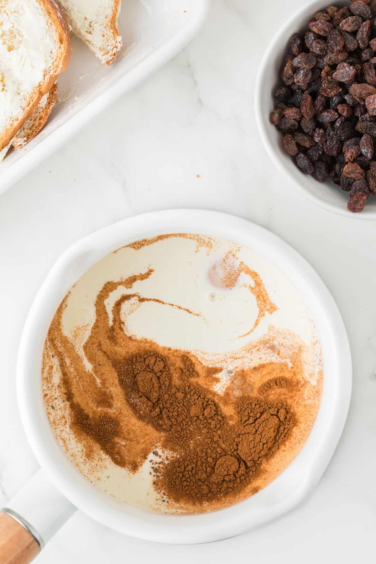 A white saucepan contains cream and a generous amount of ground cinnamon. Next to it, there is a bowl of raisins and a plate with slices of bread spread with butter or cream cheese, all on a white surface.