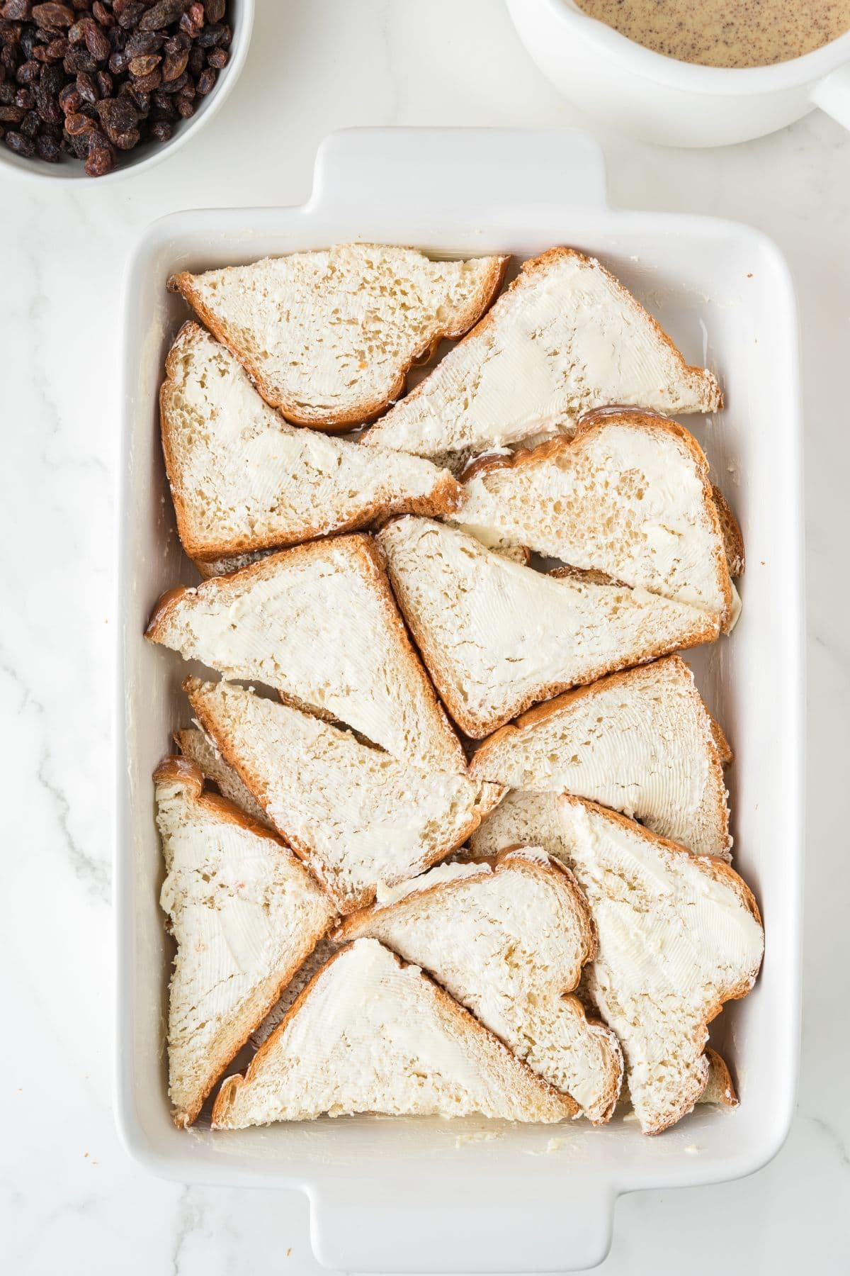 A white baking dish filled with overlapping slices of white bread spread with butter. A bowl of raisins and a container of liquid are partially visible in the background on a white surface.
