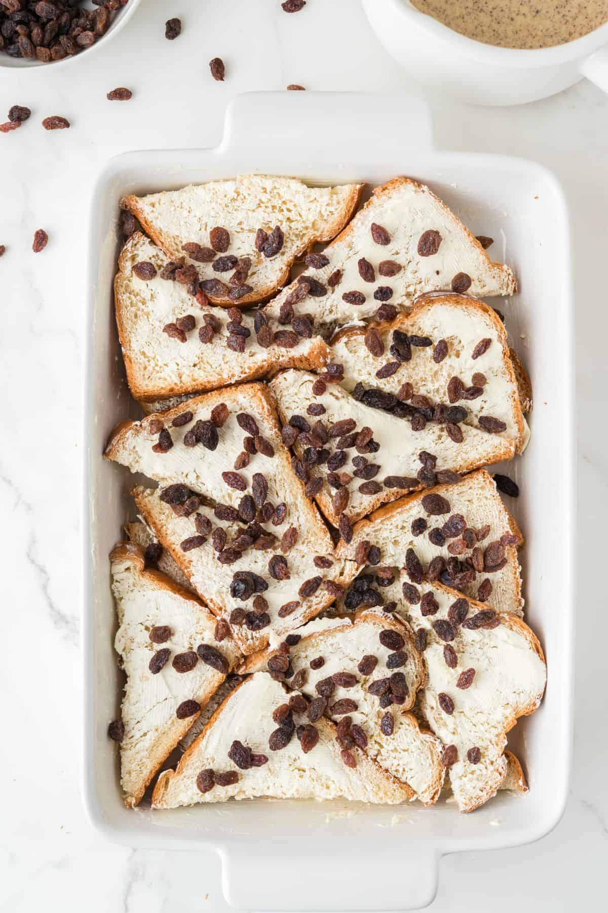 Sliced bread with butter and raisins is arranged in a white rectangular baking dish on a white surface. Some loose raisins are scattered nearby. A small bowl with a creamy mixture is partially visible in the top right corner.