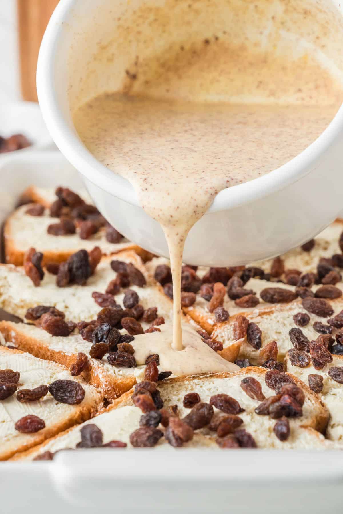 A white bowl pours a creamy egg mixture over slices of bread topped with raisins in a white baking dish, preparing a bread pudding.