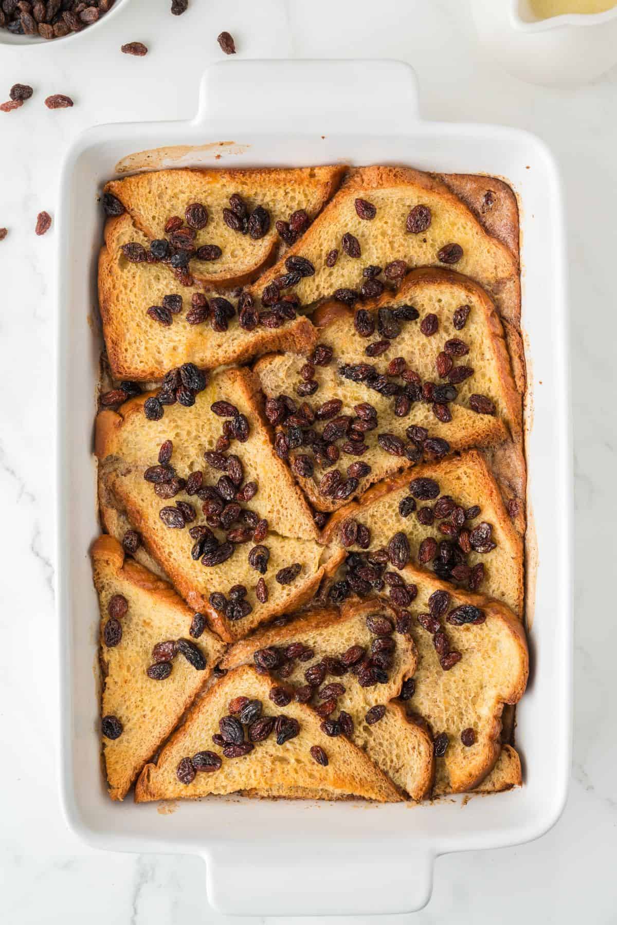 A white rectangular baking dish filled with sliced bread arranged in layers, topped with raisins, and baked to a golden brown. The dish sits on a white marble surface.