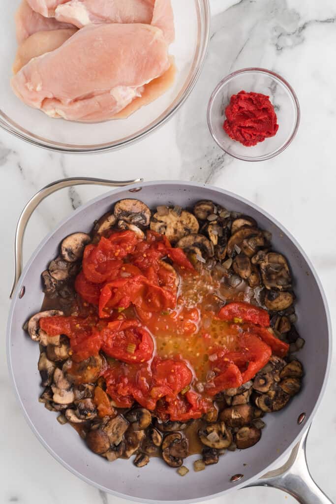 Stewed tomatoes added to mushrooms in a skillet.