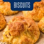 Golden brown biscuits with visible pieces of bacon and cheddar sit on parchment paper. Text on the image reads “Bacon & Cheddar Biscuits.” A honey dipper and a website URL, lanascooking.com, are also visible.