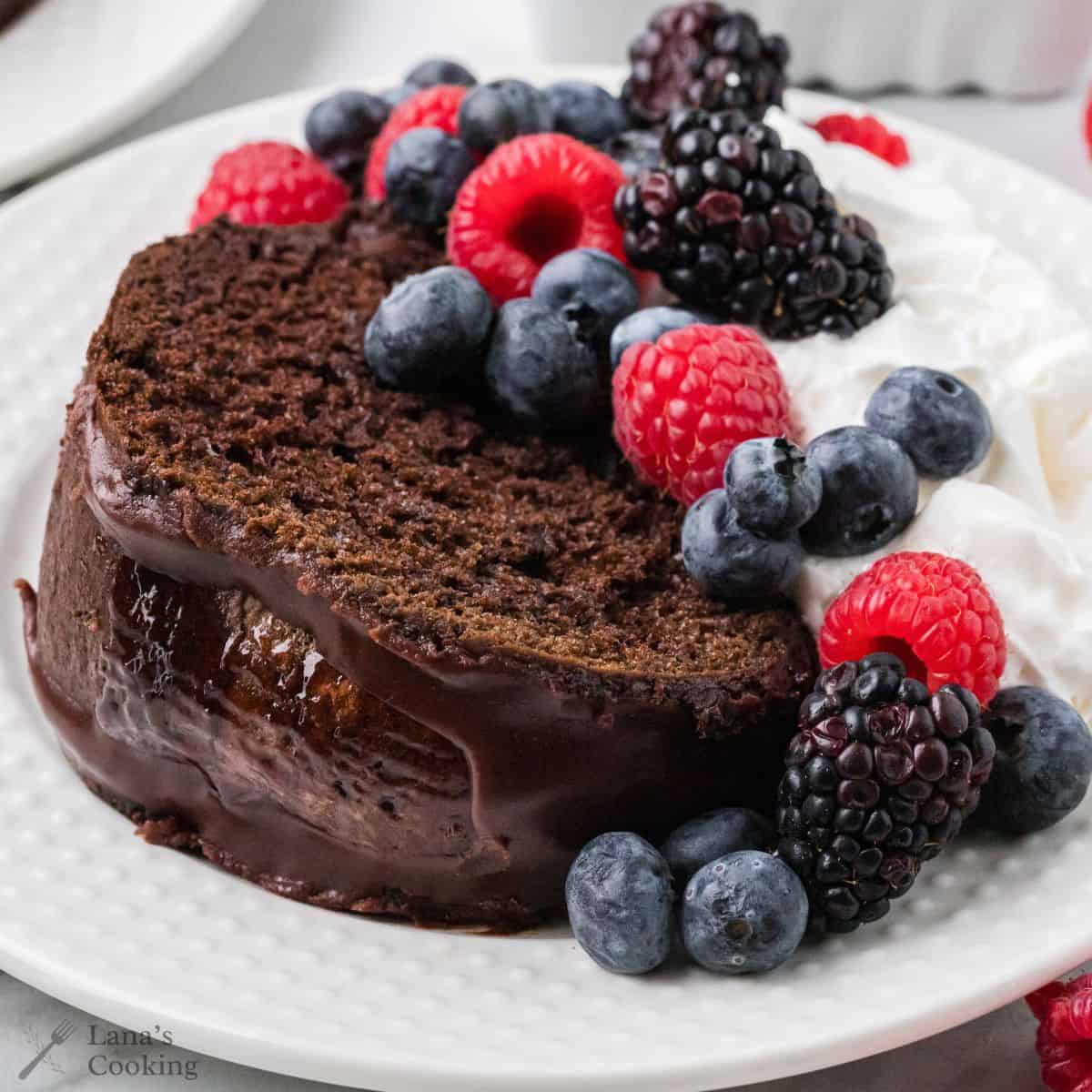 A slice of berry glazed chocolate cake with chocolate glaze, topped with fresh blueberries, raspberries, and blackberries, served with whipped cream on a white plate.