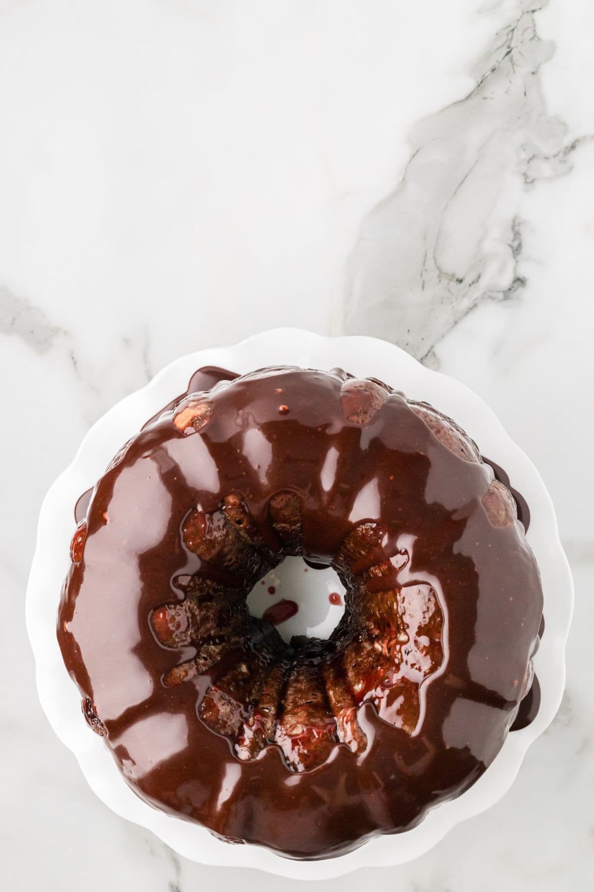 A berry glazed chocolate cake sits on a white scalloped-edge cake stand, photographed from above on a marble surface.