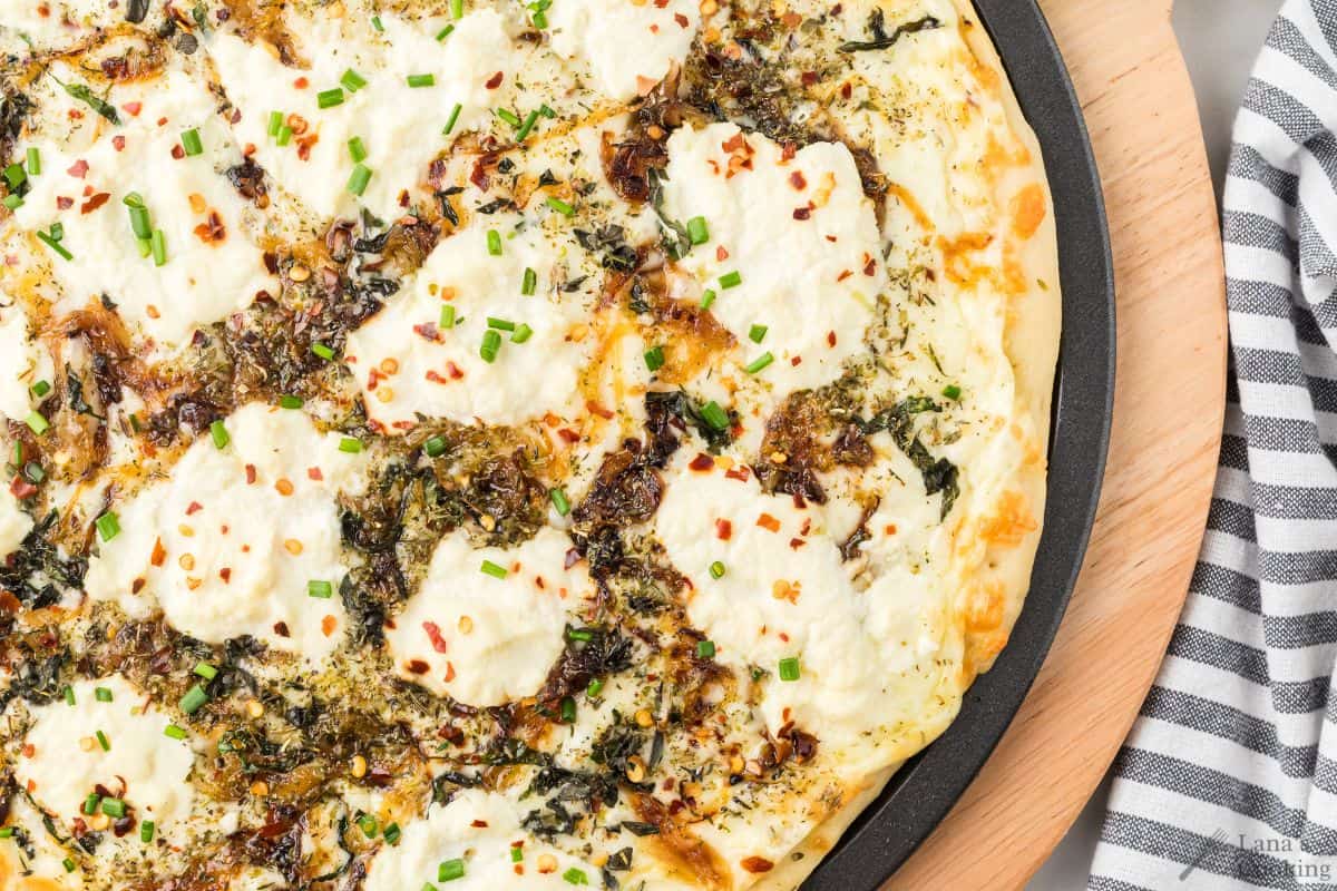 A close-up of a white pizza topped with ricotta cheese, herbs, caramelized onions, red pepper flakes, and chopped chives on a round baking tray.