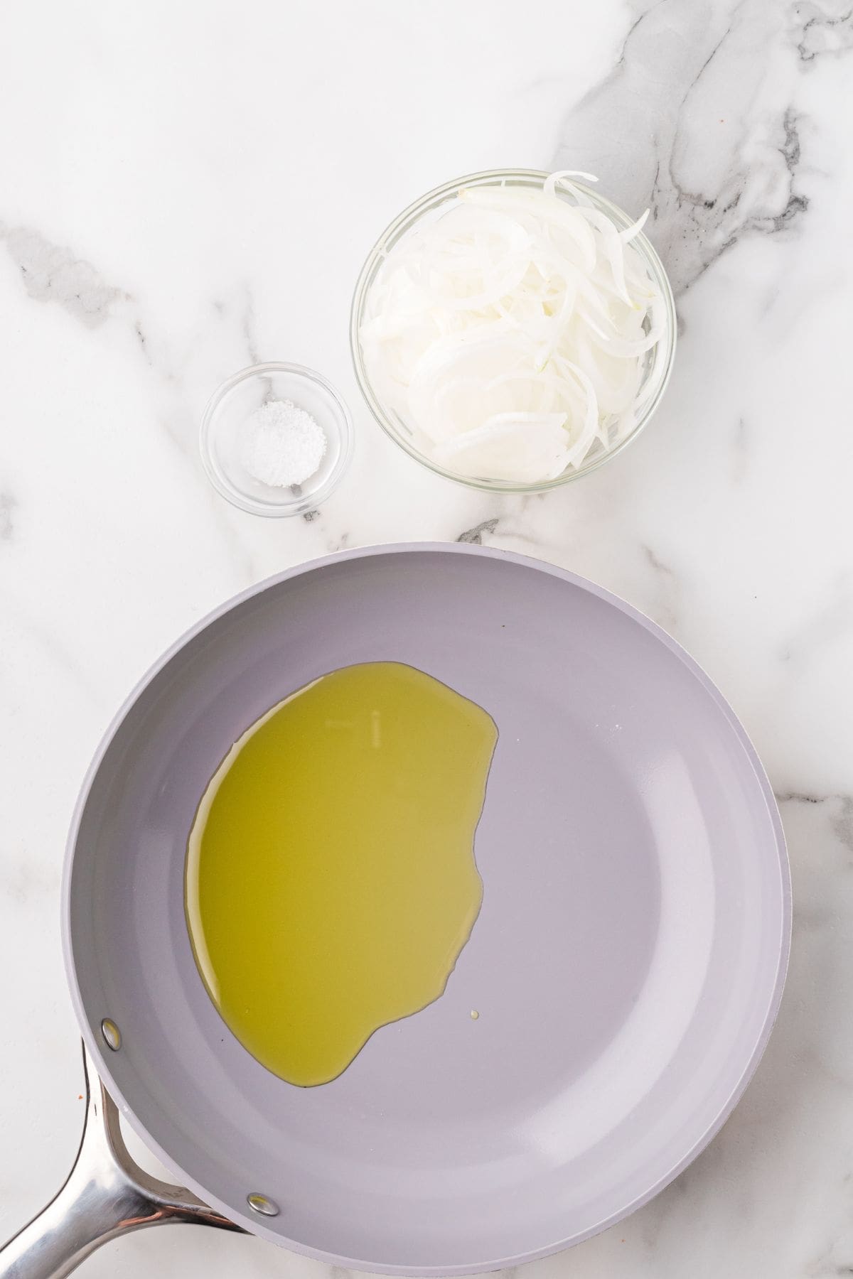 A frying pan with oil, a small bowl of salt, and a bowl of sliced onions on a white marble surface.