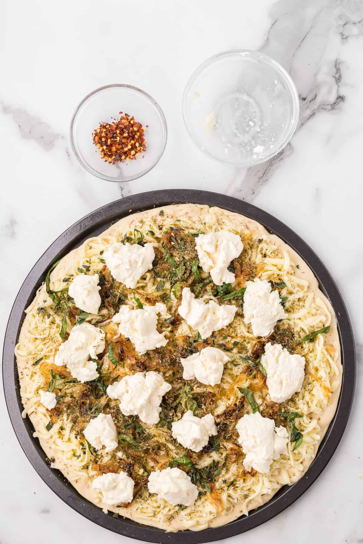 A pizza topped with cheese, herbs, and ricotta sits on a round pan. Above it are two small glass bowls, one with red pepper flakes and one empty, on a marble surface.