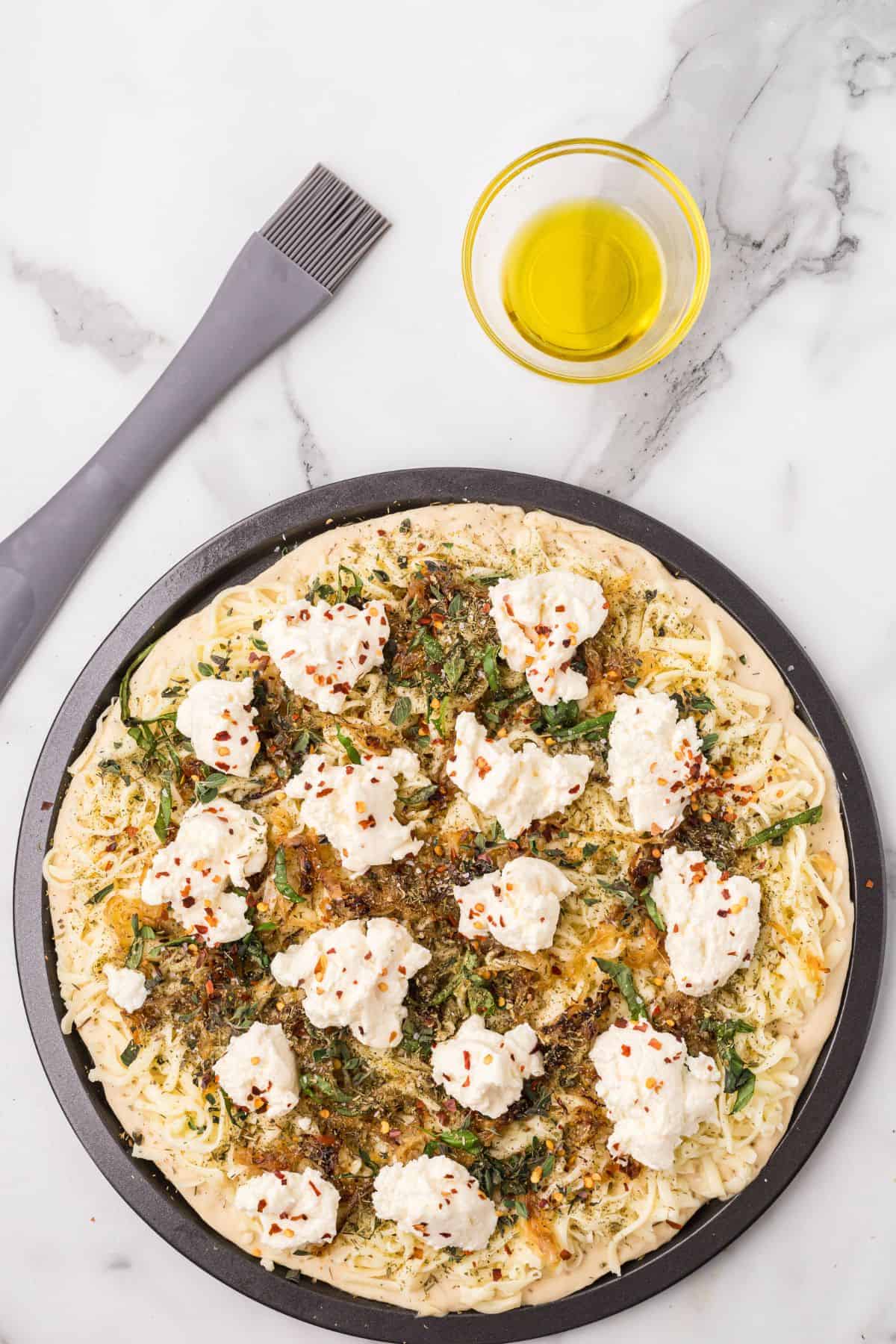 Unbaked pizza topped with cheese, herbs, and red pepper flakes, on a pizza pan next to a bowl of olive oil and a pastry brush on a marble surface.