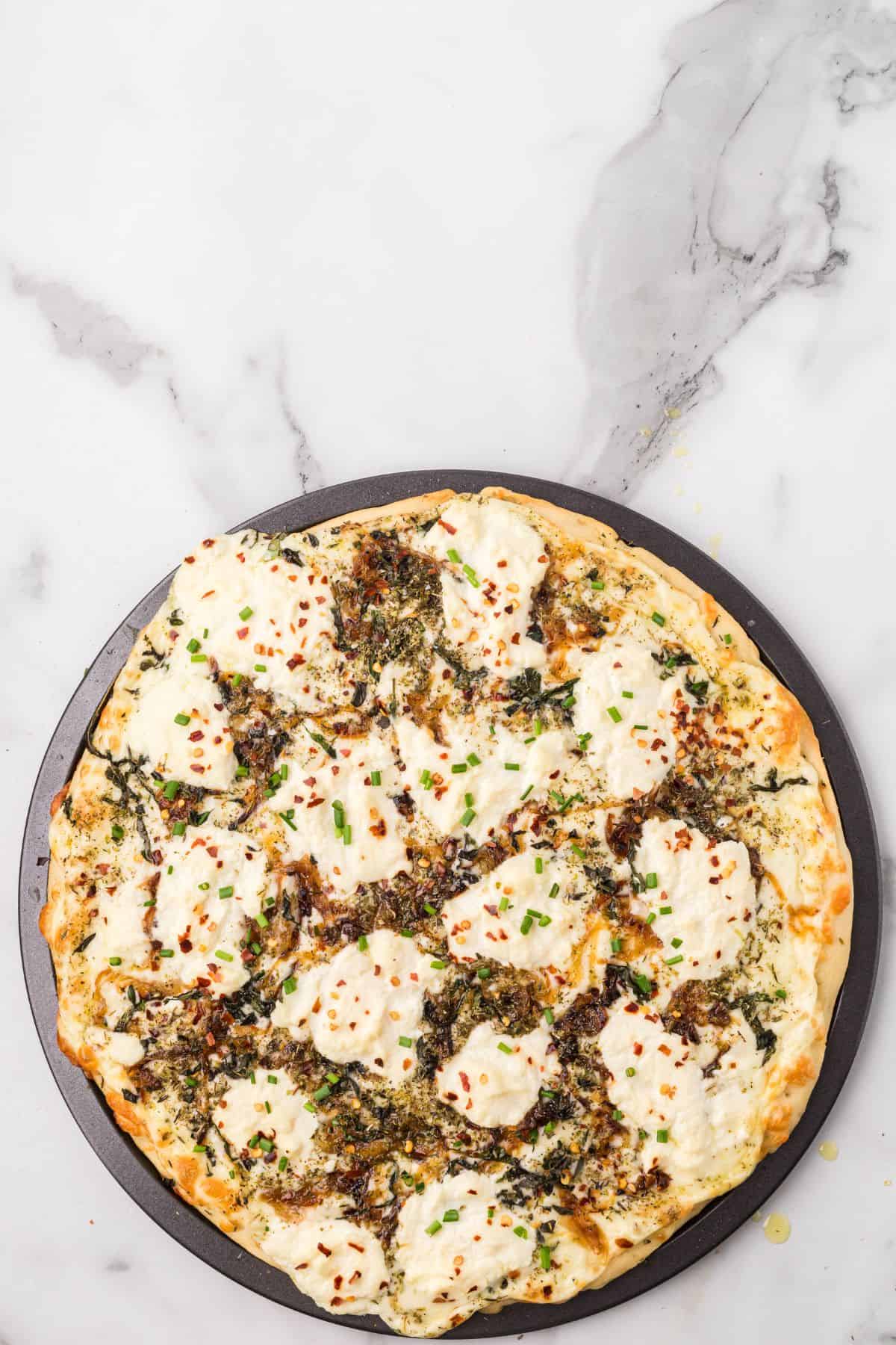 A baked pizza topped with dollops of ricotta cheese, herbs, and red pepper flakes on a round tray, set on a marble surface.