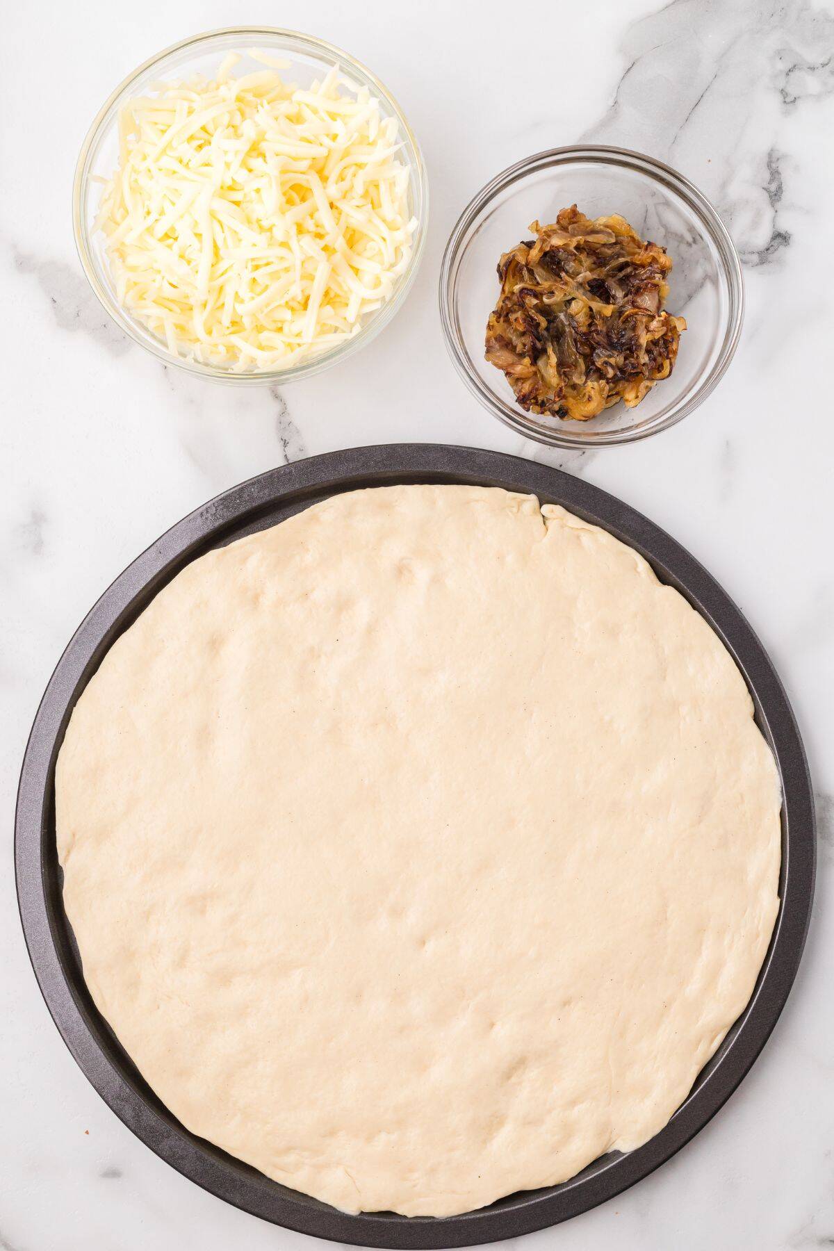A round pizza dough on a baking pan, with bowls of shredded cheese and caramelized onions on a marble surface.