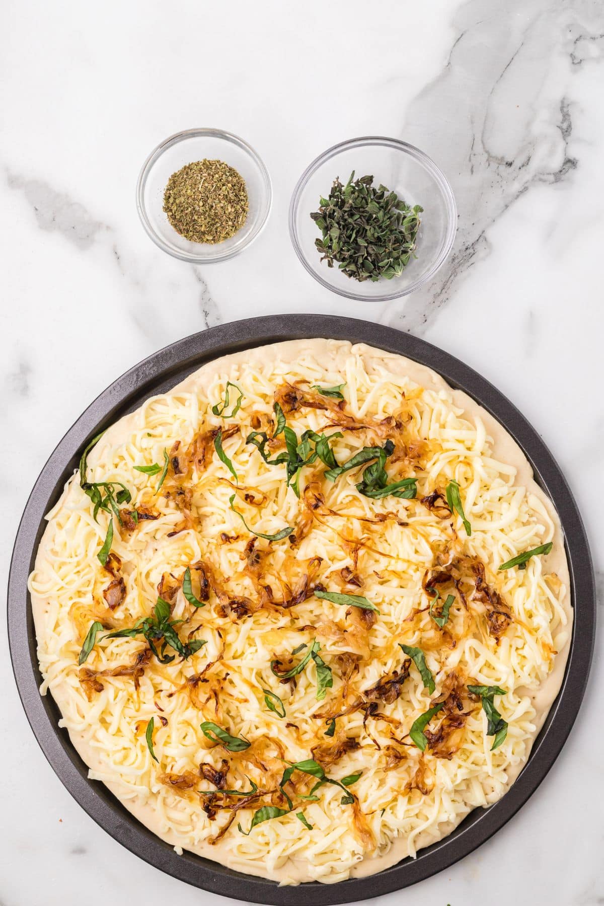 Unbaked pizza topped with shredded cheese, caramelized onions, and fresh basil on a round pan, with small bowls of dried and fresh herbs on a marble surface.