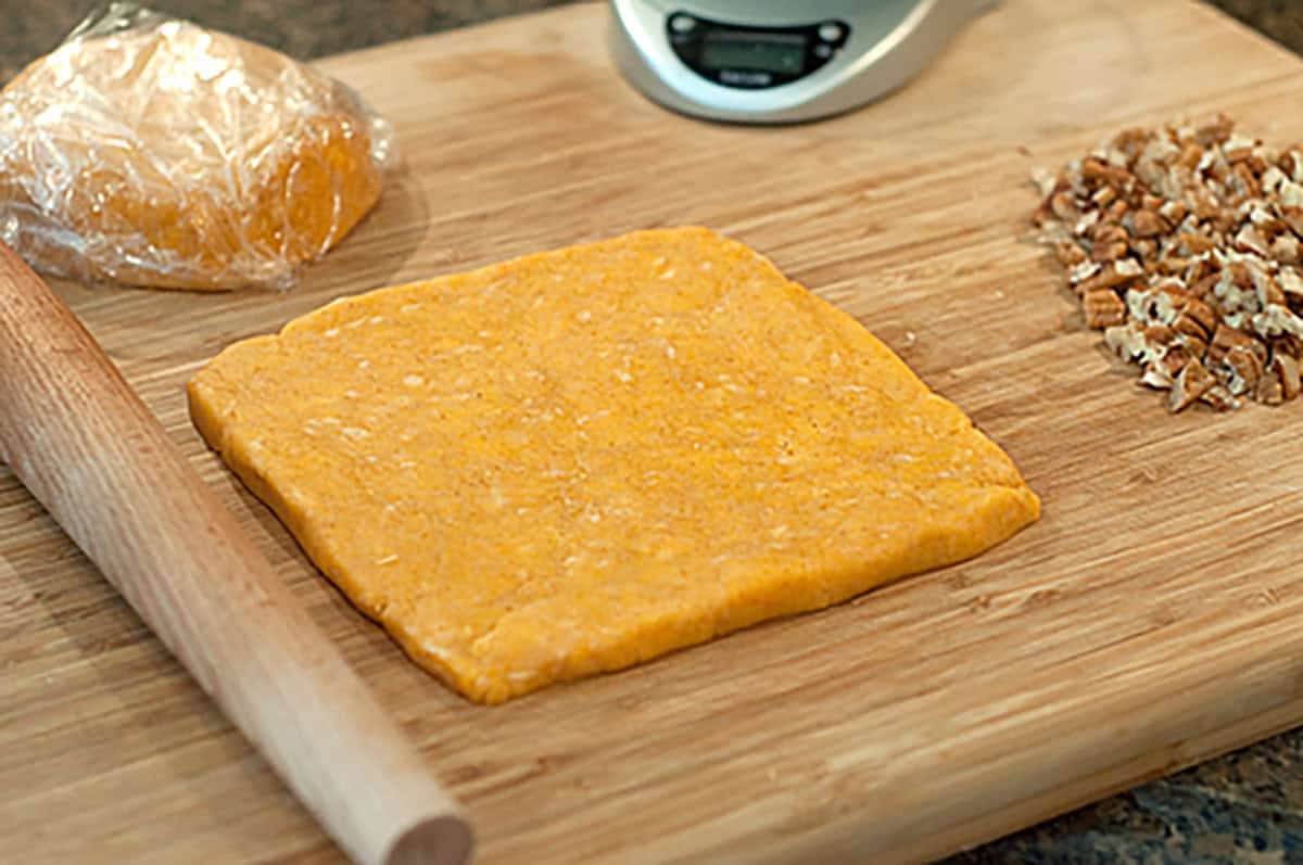 A square slab of orange dough, a rolling pin, chopped nuts, a digital scale, and plastic-wrapped dough on a wooden cutting board.