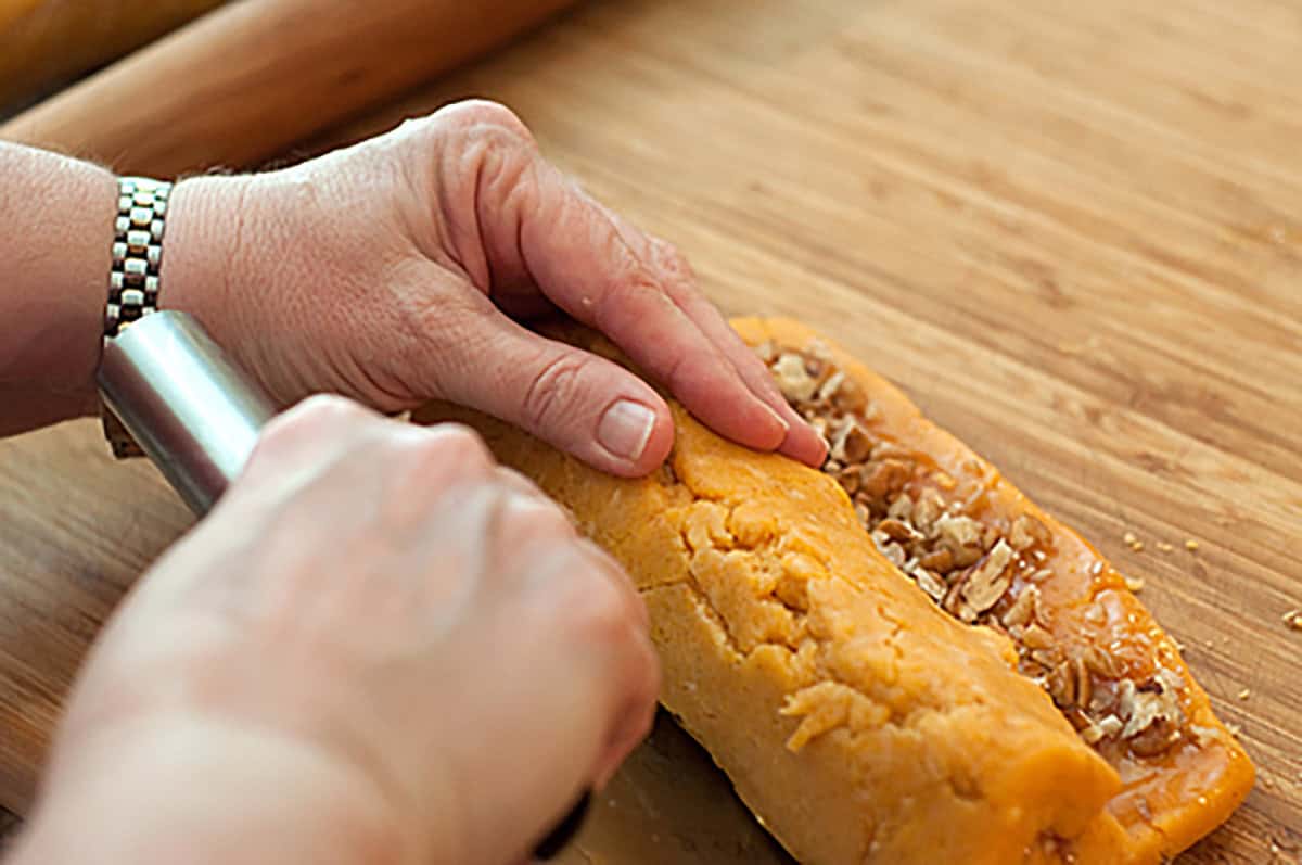 A person&rsquo;s hands rolling dough with a nut filling on a wooden surface, using a metal tool to shape the dough.